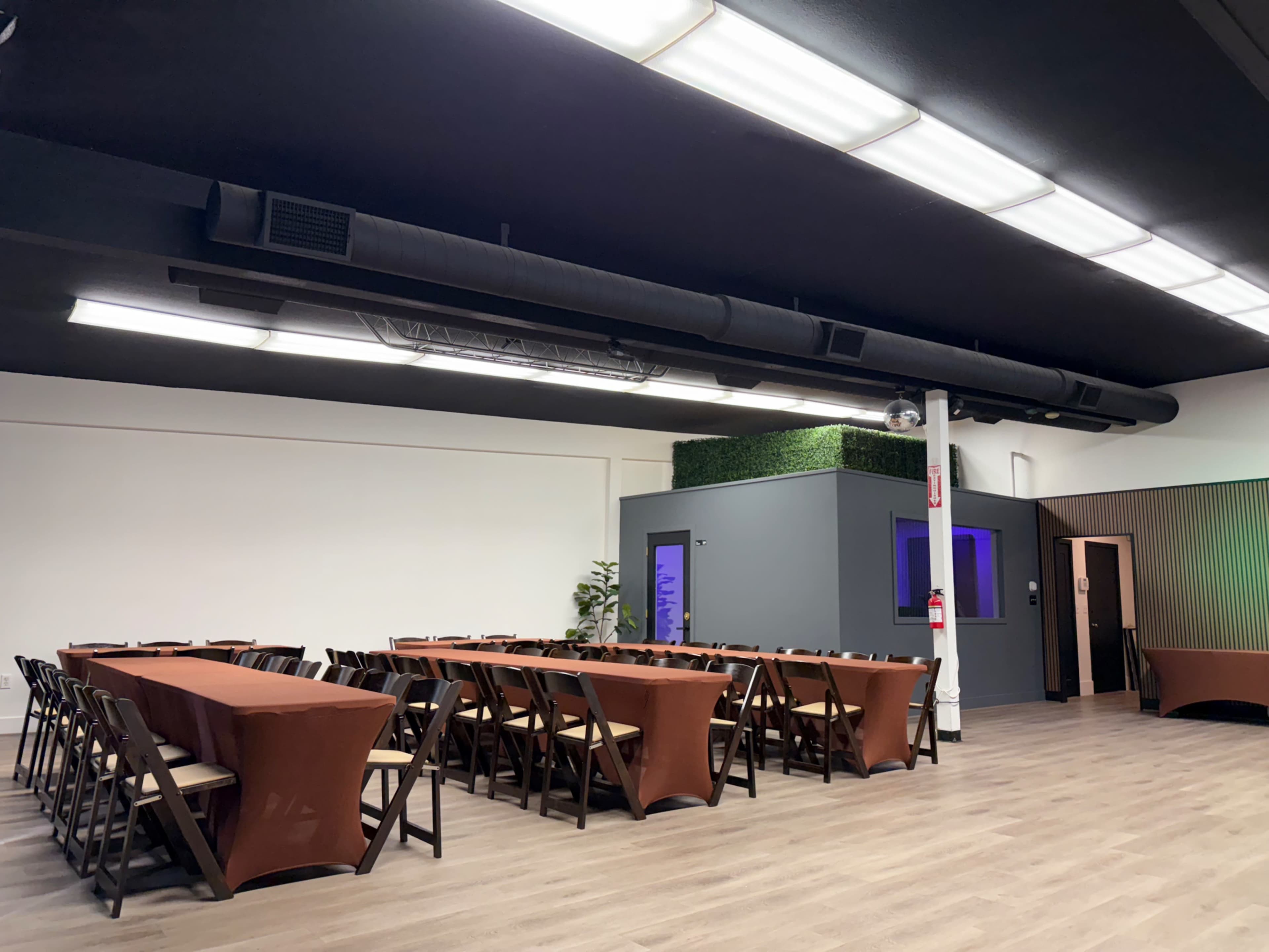 The image shows a spacious event room with multiple tables arranged in a U-shape and a grey structure in the background, illuminated by colored lights.