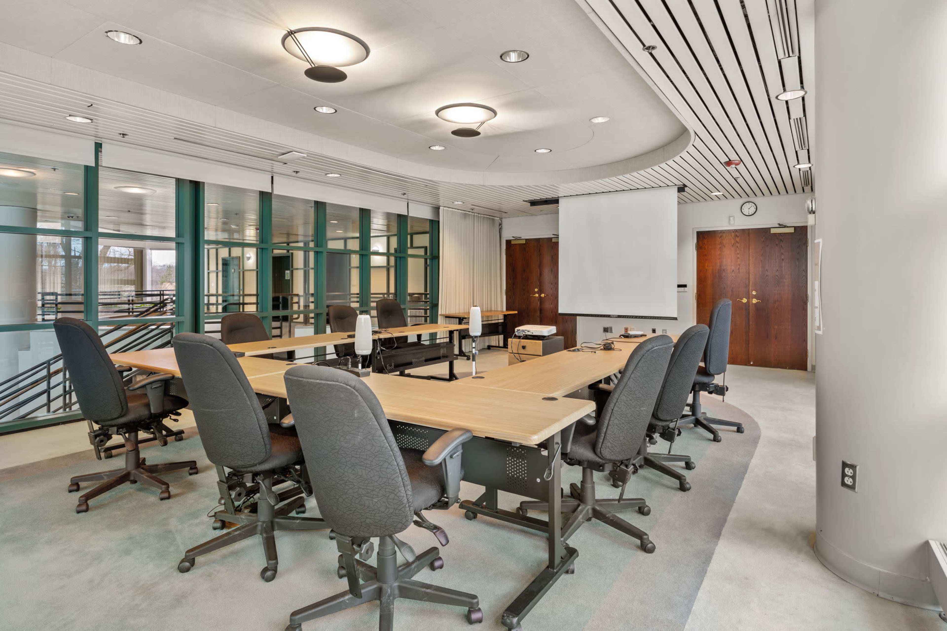 A meeting room features a large, oval table surrounded by ergonomic chairs, with a projector screen at one end and large windows along the walls.