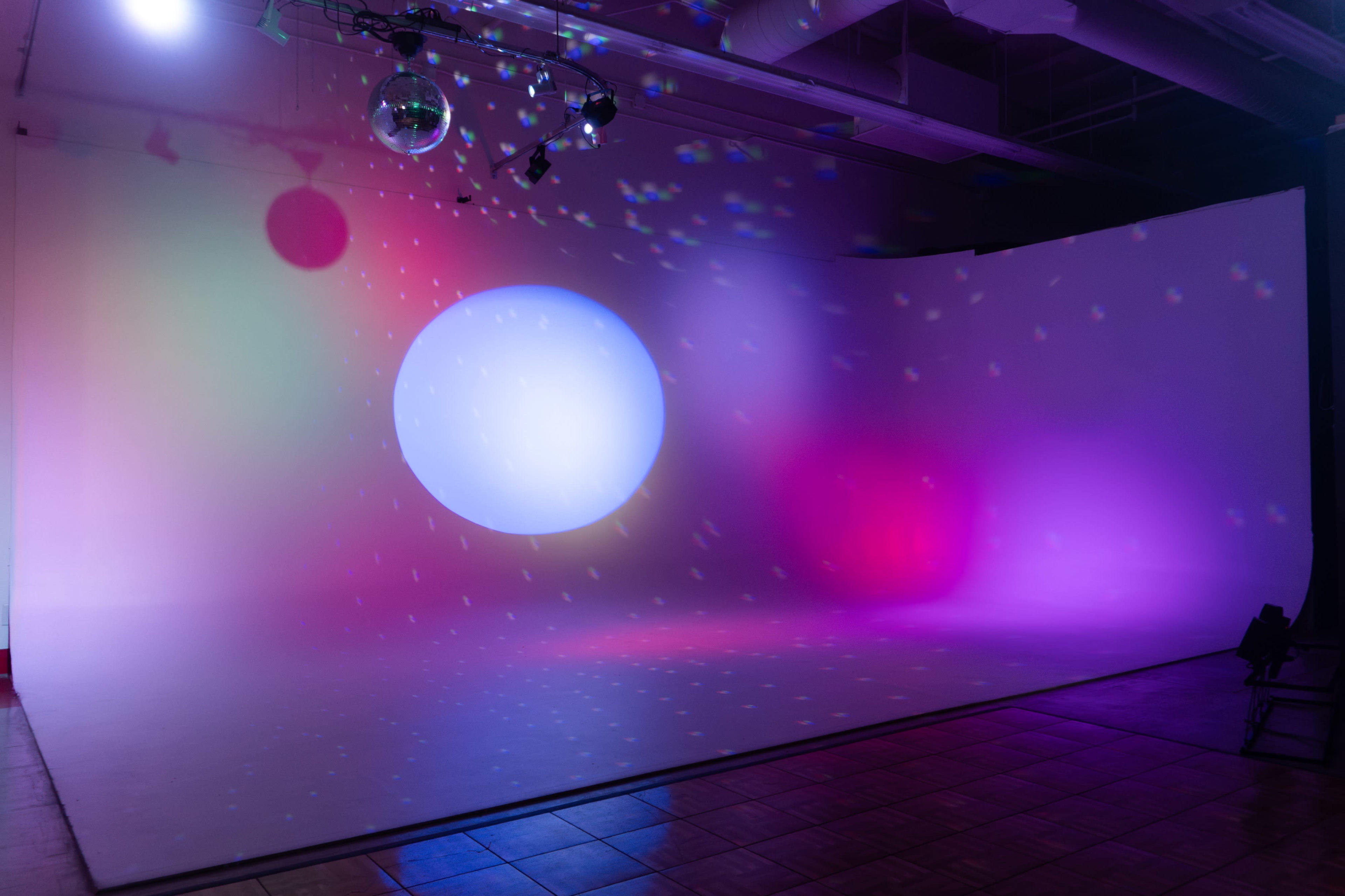 The image shows a studio space with a curved white backdrop illuminated by colorful lights and reflections from disco balls.