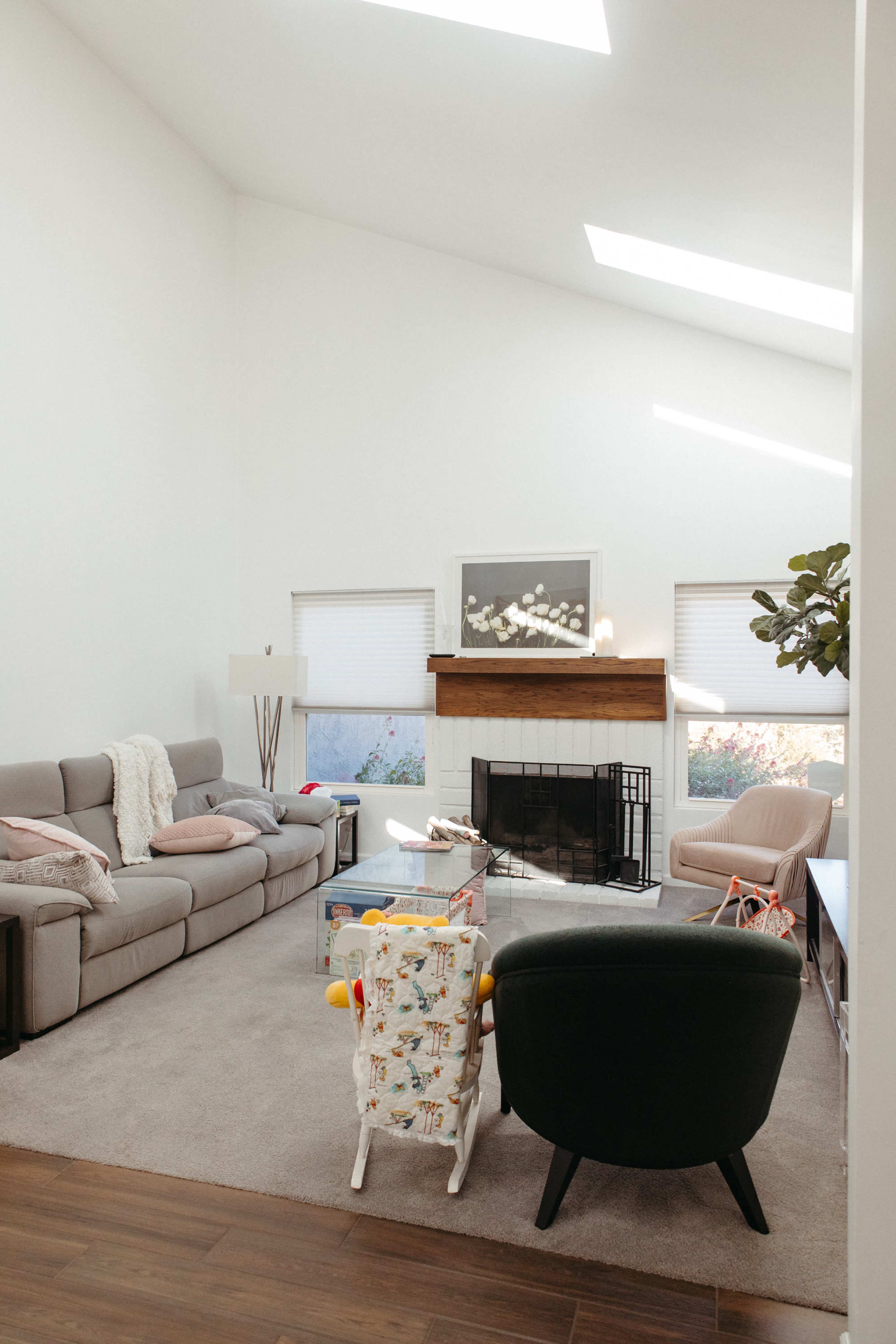 A bright and airy living room features a gray sectional sofa, a wooden fireplace with a mantel, and a small children’s chair, all illuminated by natural light from skylights.