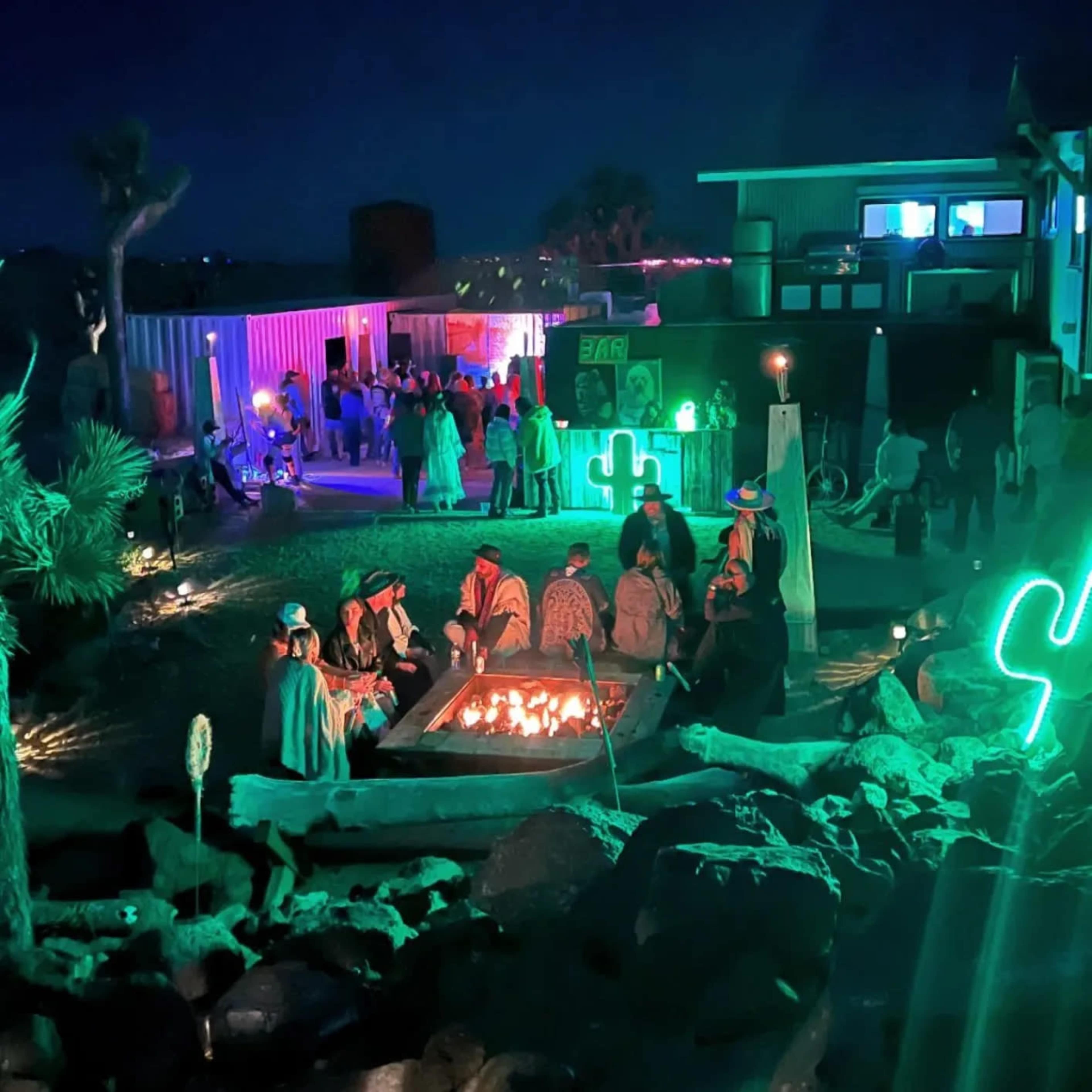 A group of people gathers around a fire pit in a desert setting at night, with a modern building and greenery in the background.