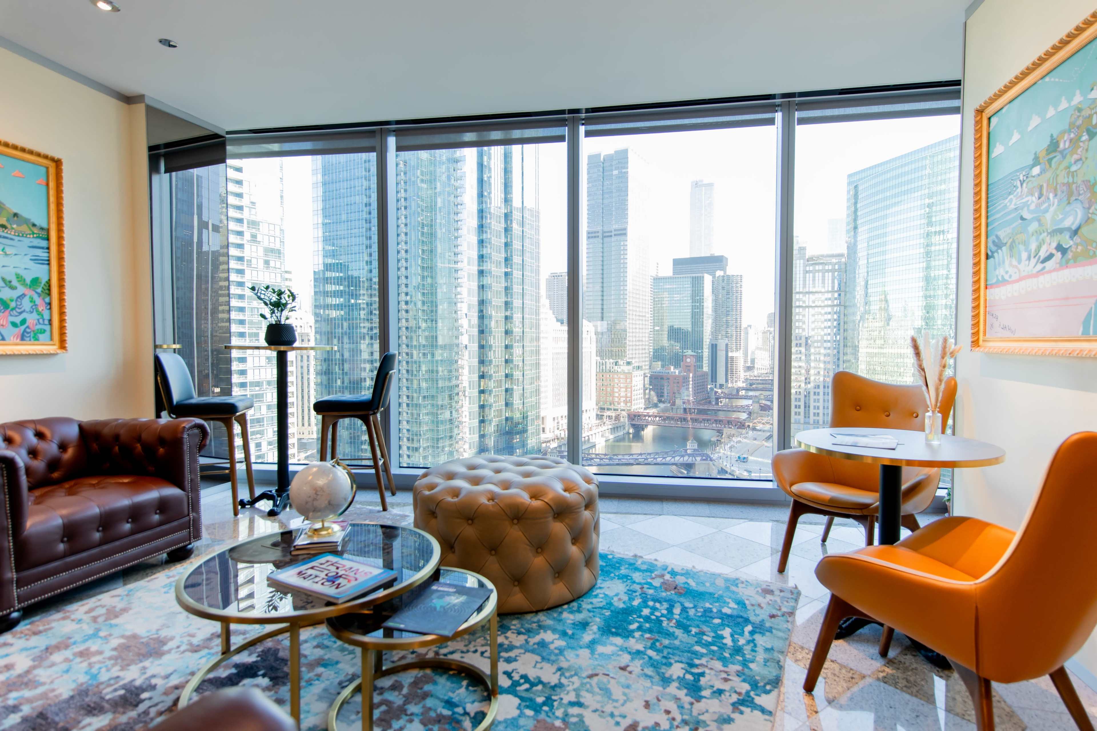 A modern room features a glass wall with a city view, plush seating, and a round table.
