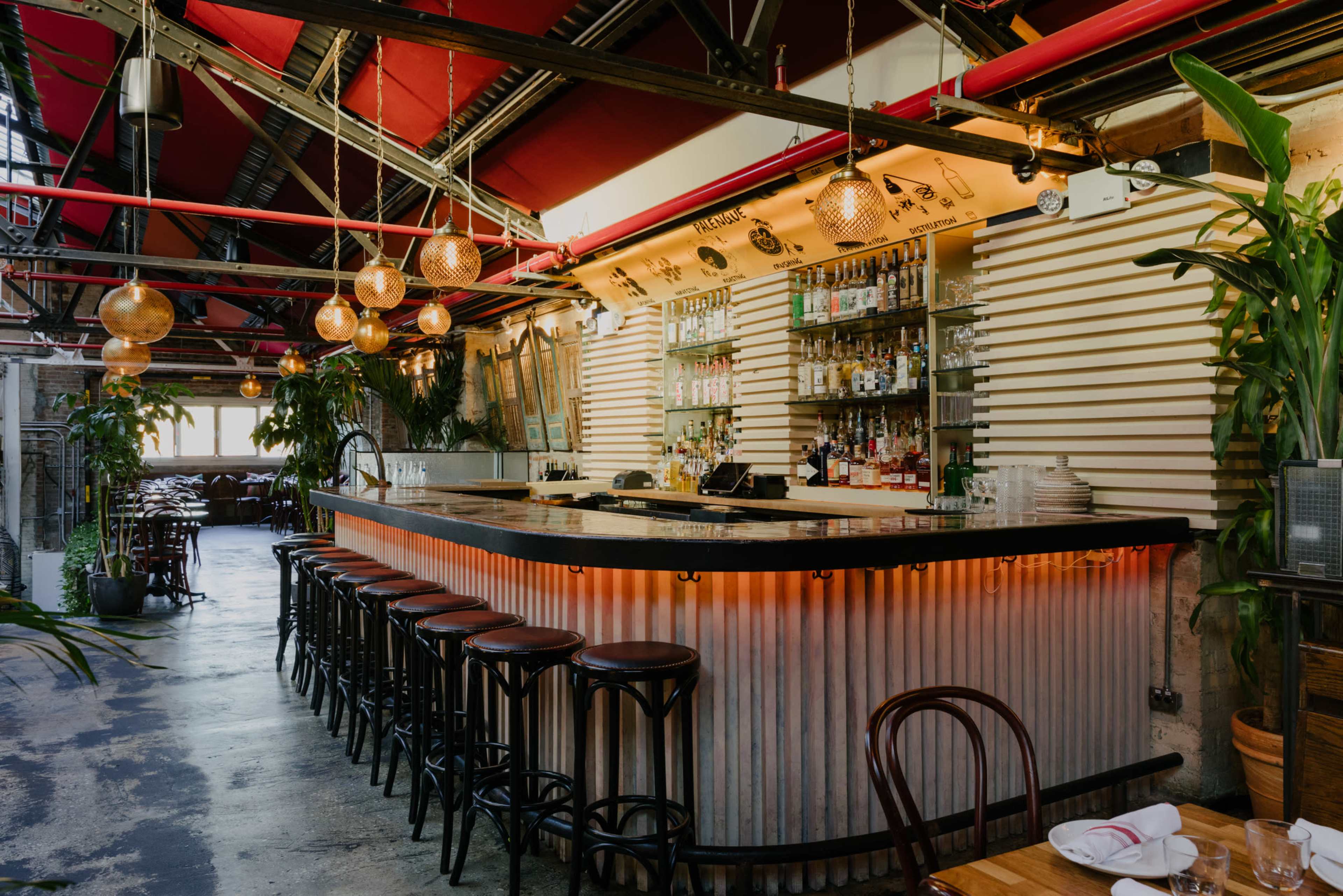 A modern bar with a curved countertop, surrounded by stools, and illuminated by pendant lights, in a spacious venue with plants and wooden furniture.