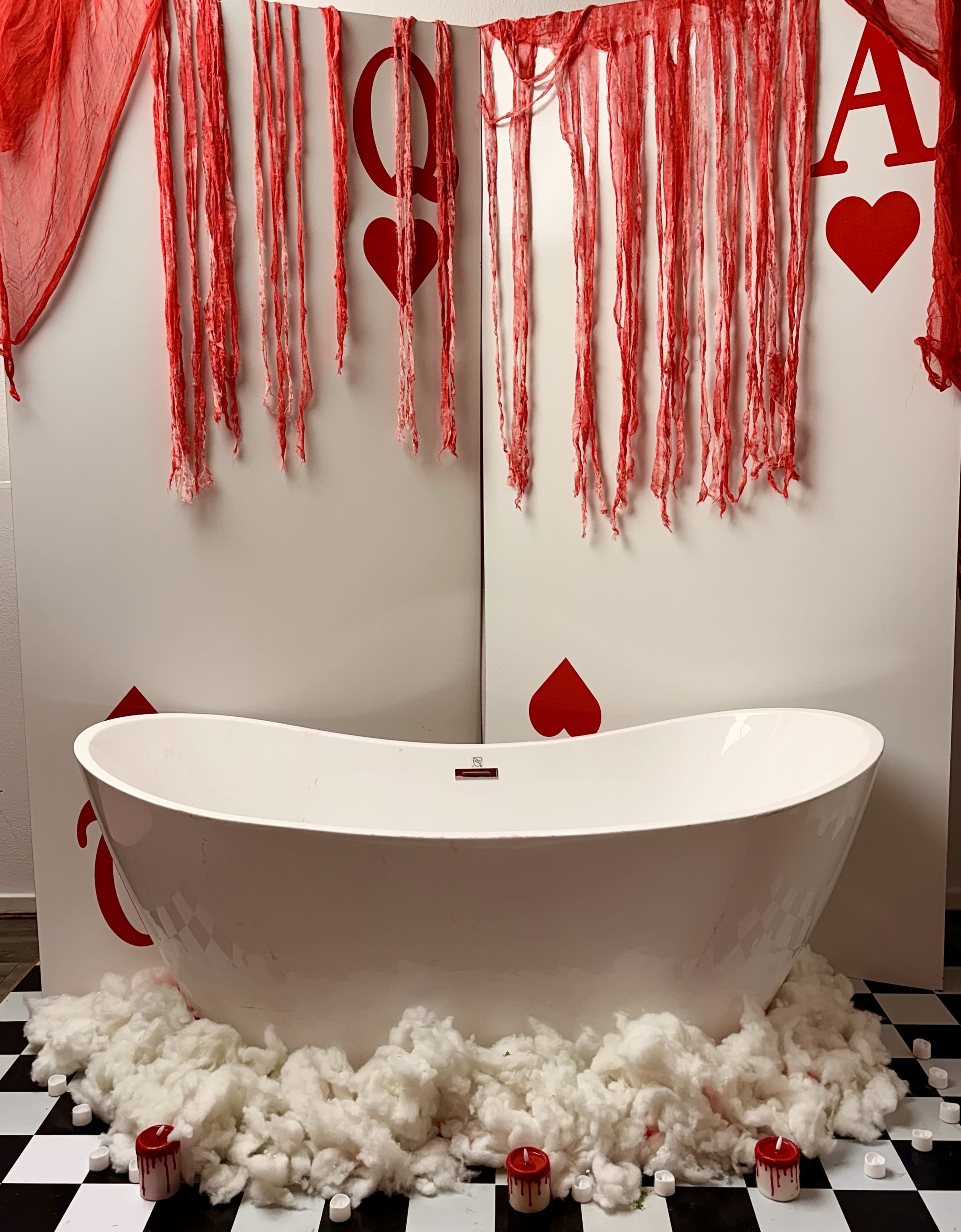 A freestanding white bathtub is seated on a checkerboard floor surrounded by cotton-like material and decorated with red streamers and large playing card motifs.