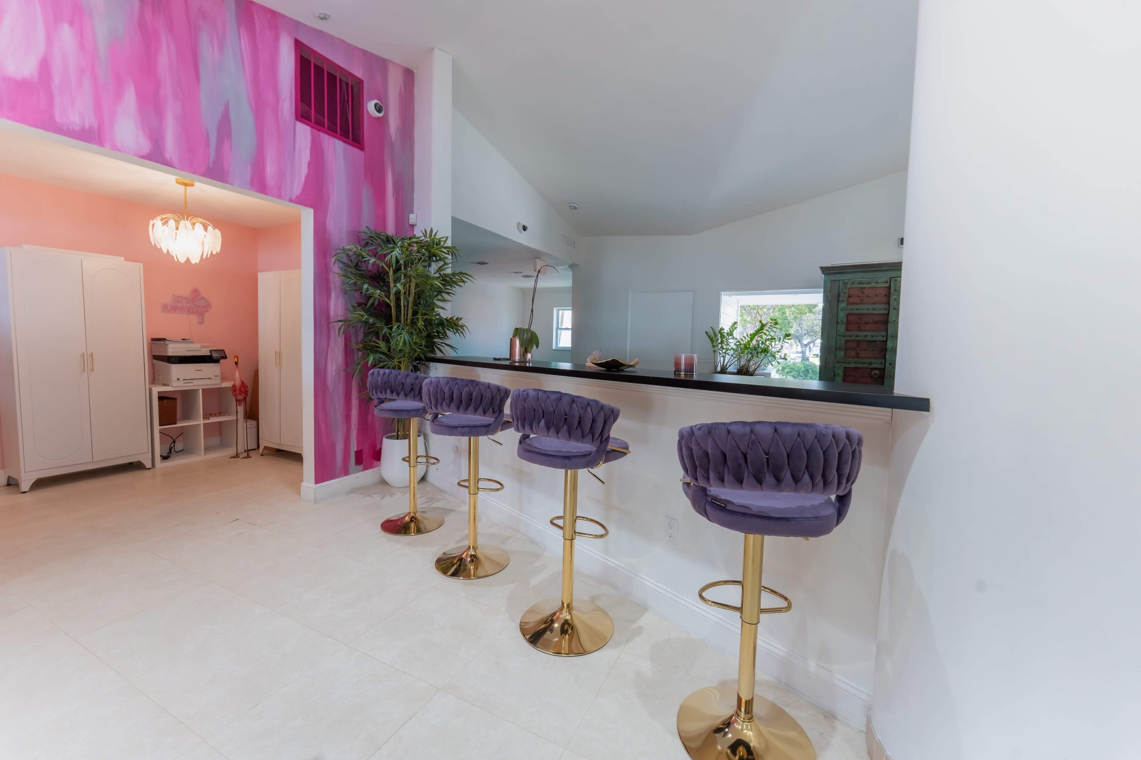 The image shows a modern bar area with four purple velvet stools and a pink and white painted wall, complemented by a light-colored tile floor.