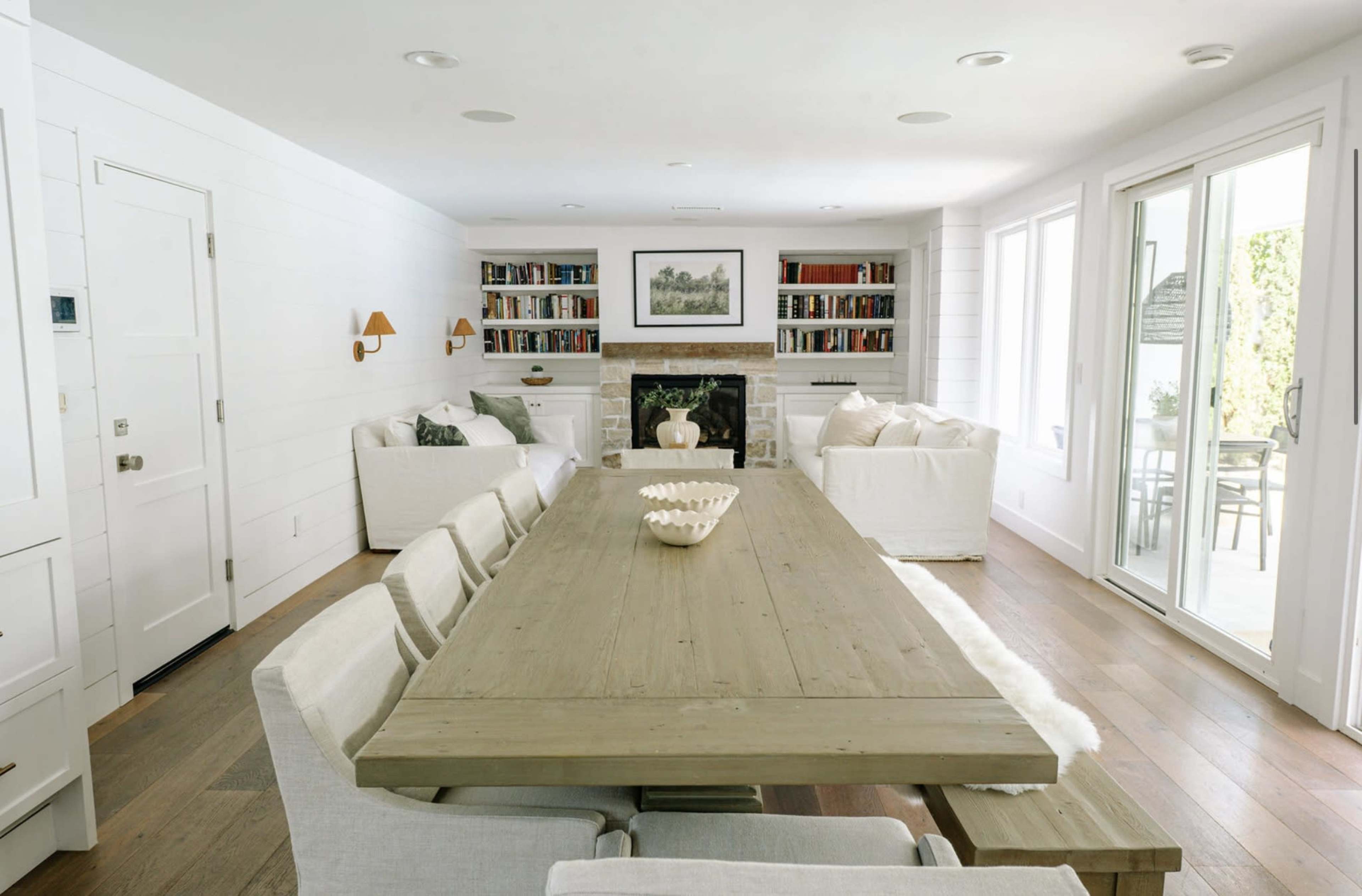 A spacious living room features a large wooden dining table at the center, surrounded by upholstered chairs, with a fireplace and bookshelves in the background.