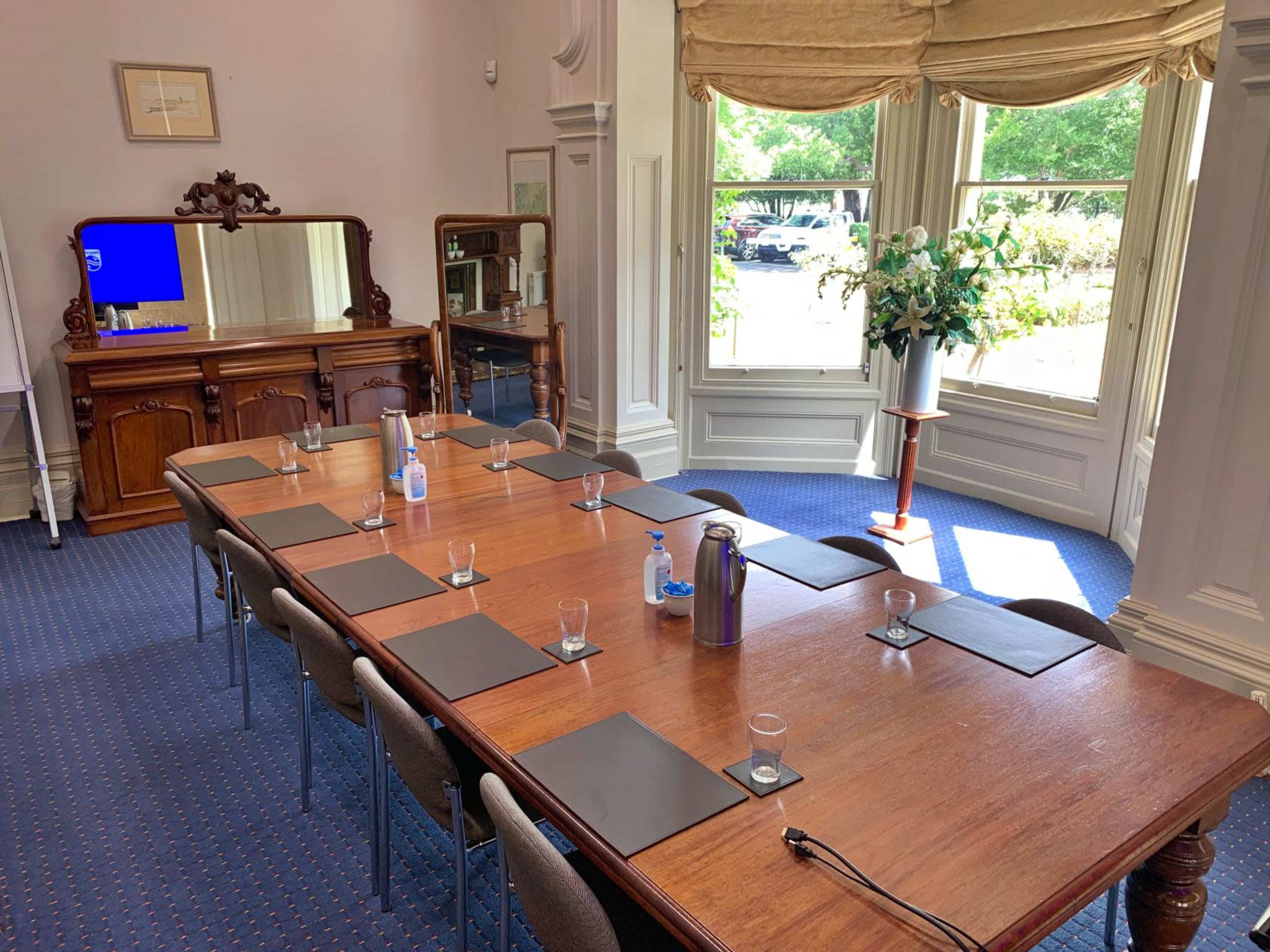 A conference room features a long wooden table surrounded by chairs, with a sideboard and large windows allowing natural light to enter.