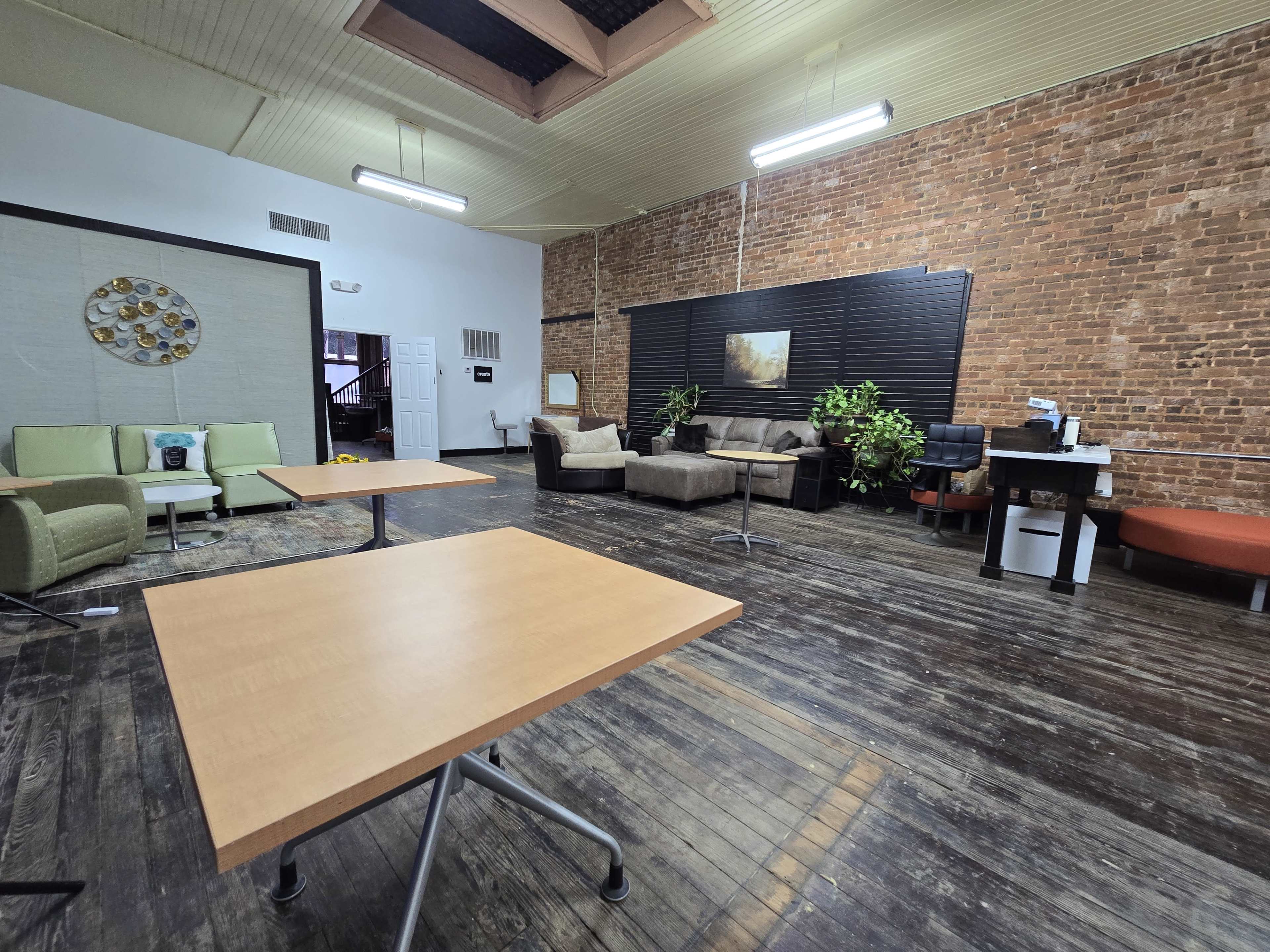 The image shows a spacious room with wooden floors, exposed brick walls, and a mix of seating areas including sofas, chairs, and tables.