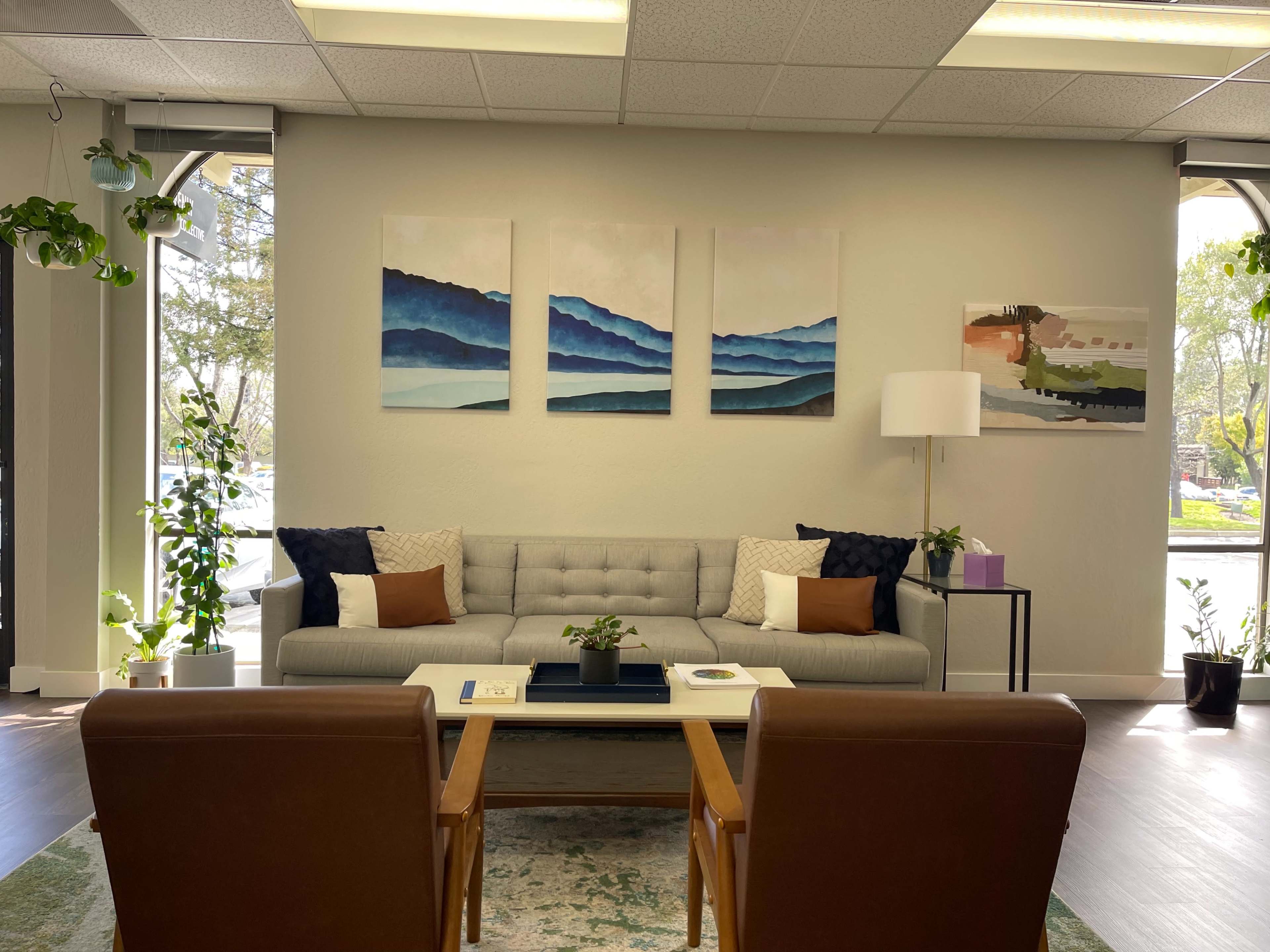 A cozy waiting area with a gray sofa, two brown chairs, a coffee table, and colorful landscape paintings on the wall.