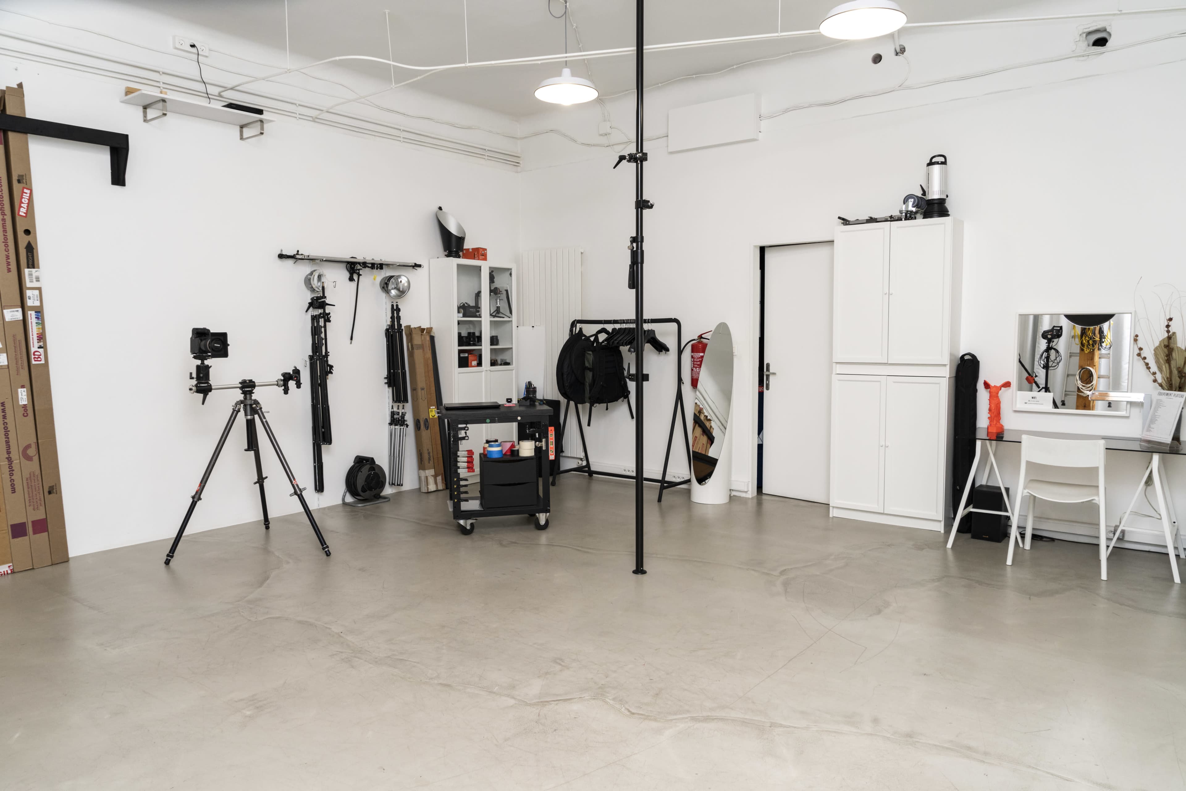 A minimalist studio space features a tripod-mounted camera, a portable cart with equipment, and various backdrops against white walls.