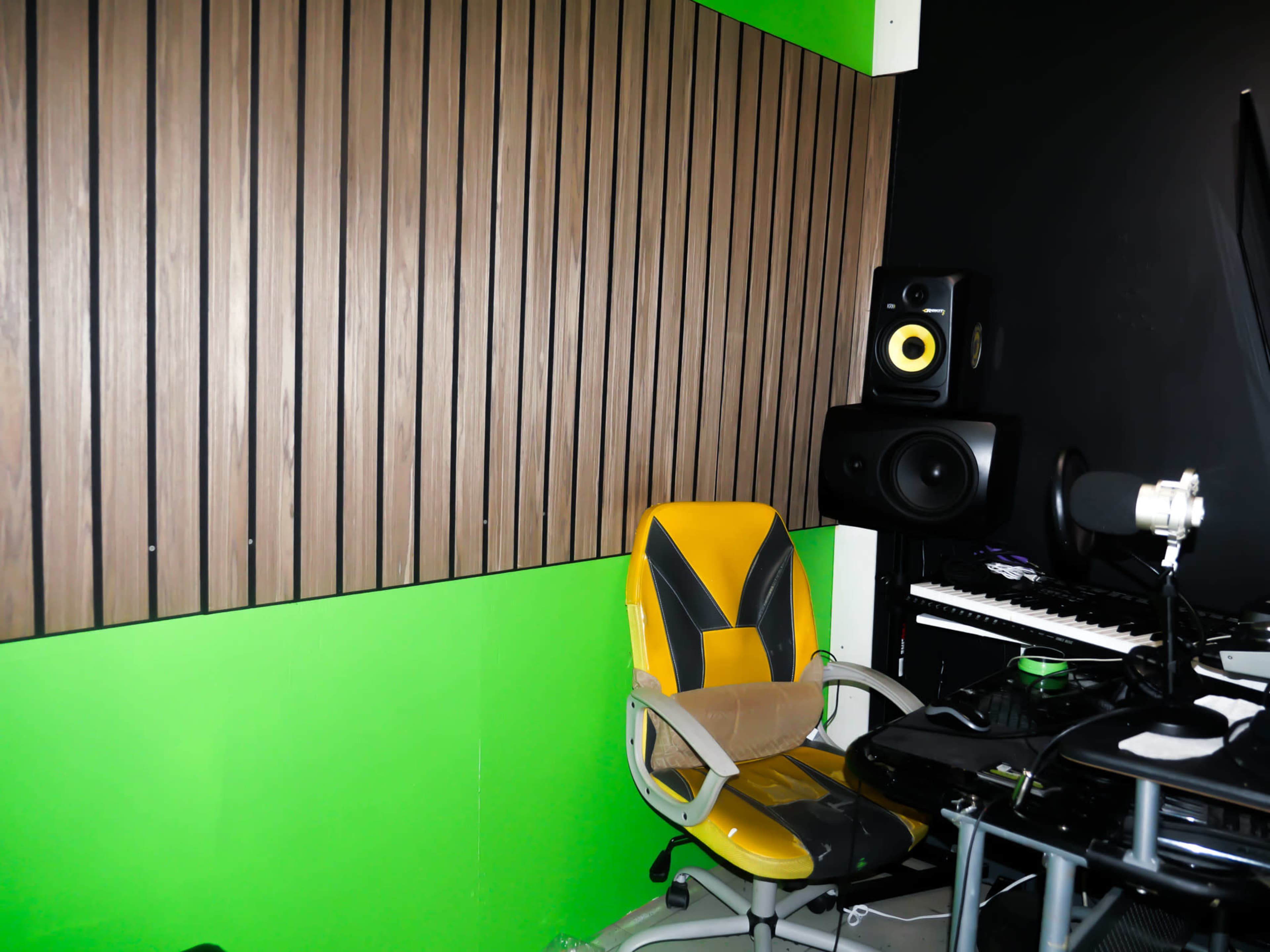 The image shows a music studio corner featuring a green wall, wooden paneling, a microphone, a keyboard, and a yellow office chair.