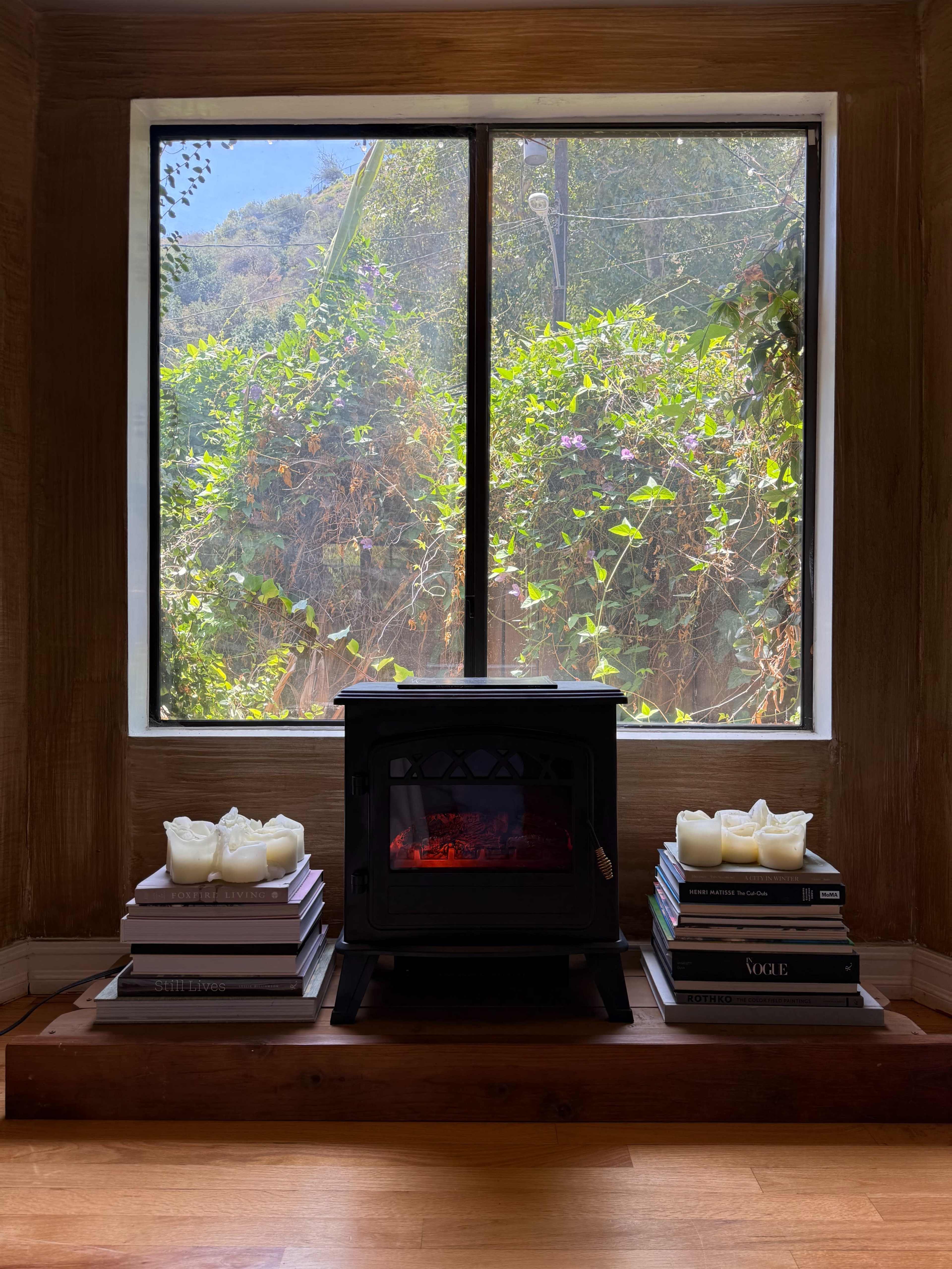 A small black wood stove sits on a wooden platform between stacks of books, with a view of greenery visible through a large window behind it.