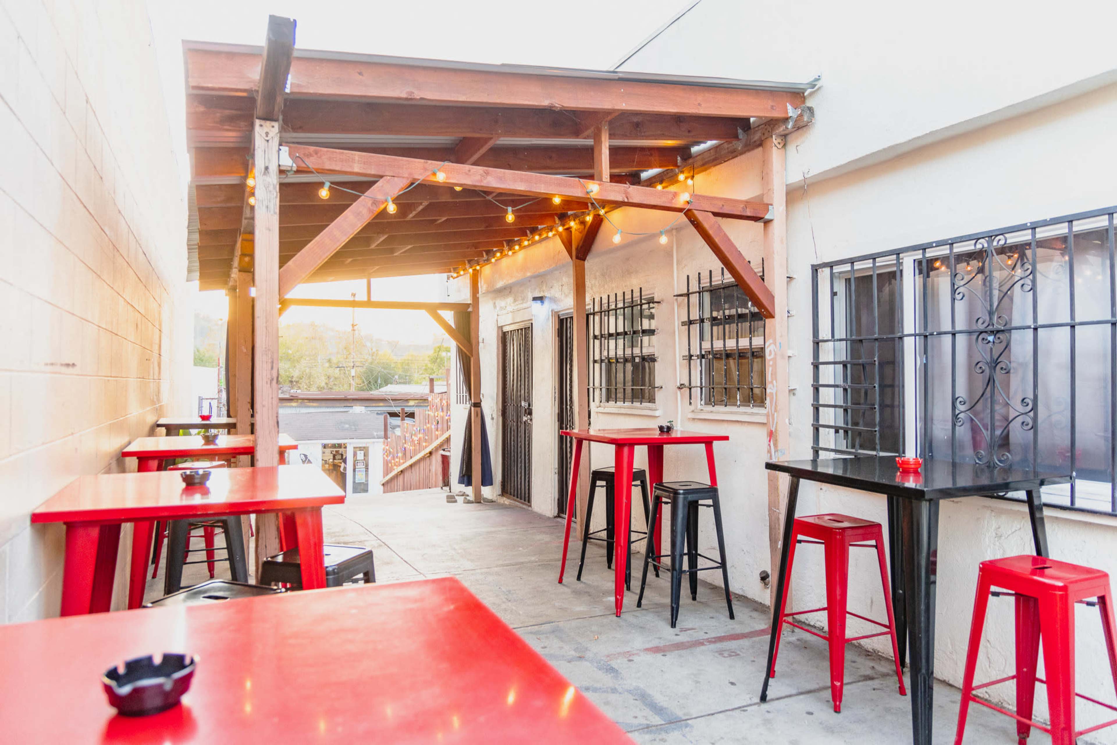 A patio area with wooden beams and string lights, featuring red and black bar tables and stools.