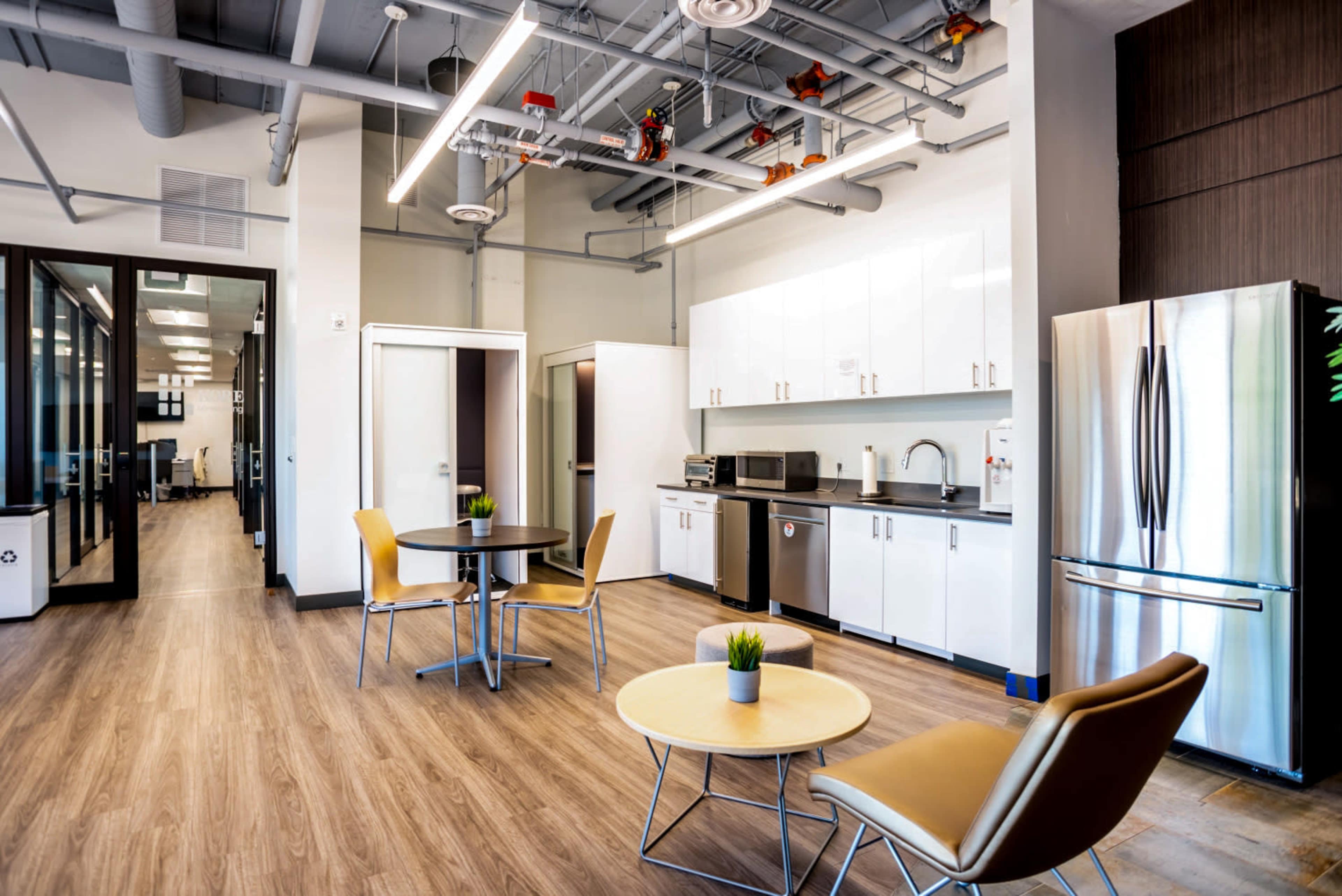 The image shows a modern break room with a kitchenette, featuring a table and chairs, stainless steel appliances, and a clean, minimalist design.