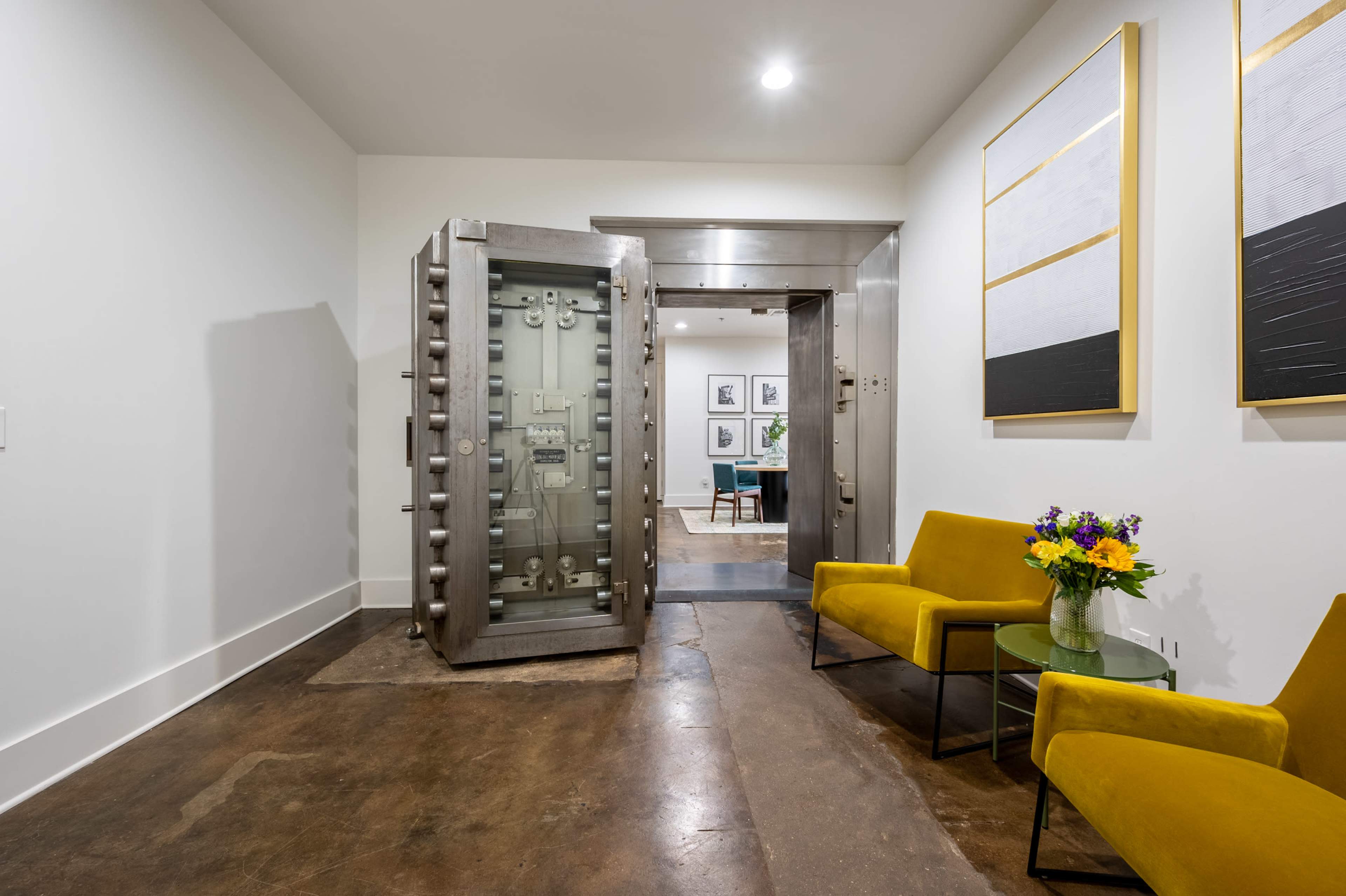 A large metallic vault door stands open in a modern hallway, revealing a room with a table and chairs, while two yellow chairs and a vase of flowers occupy the foreground.