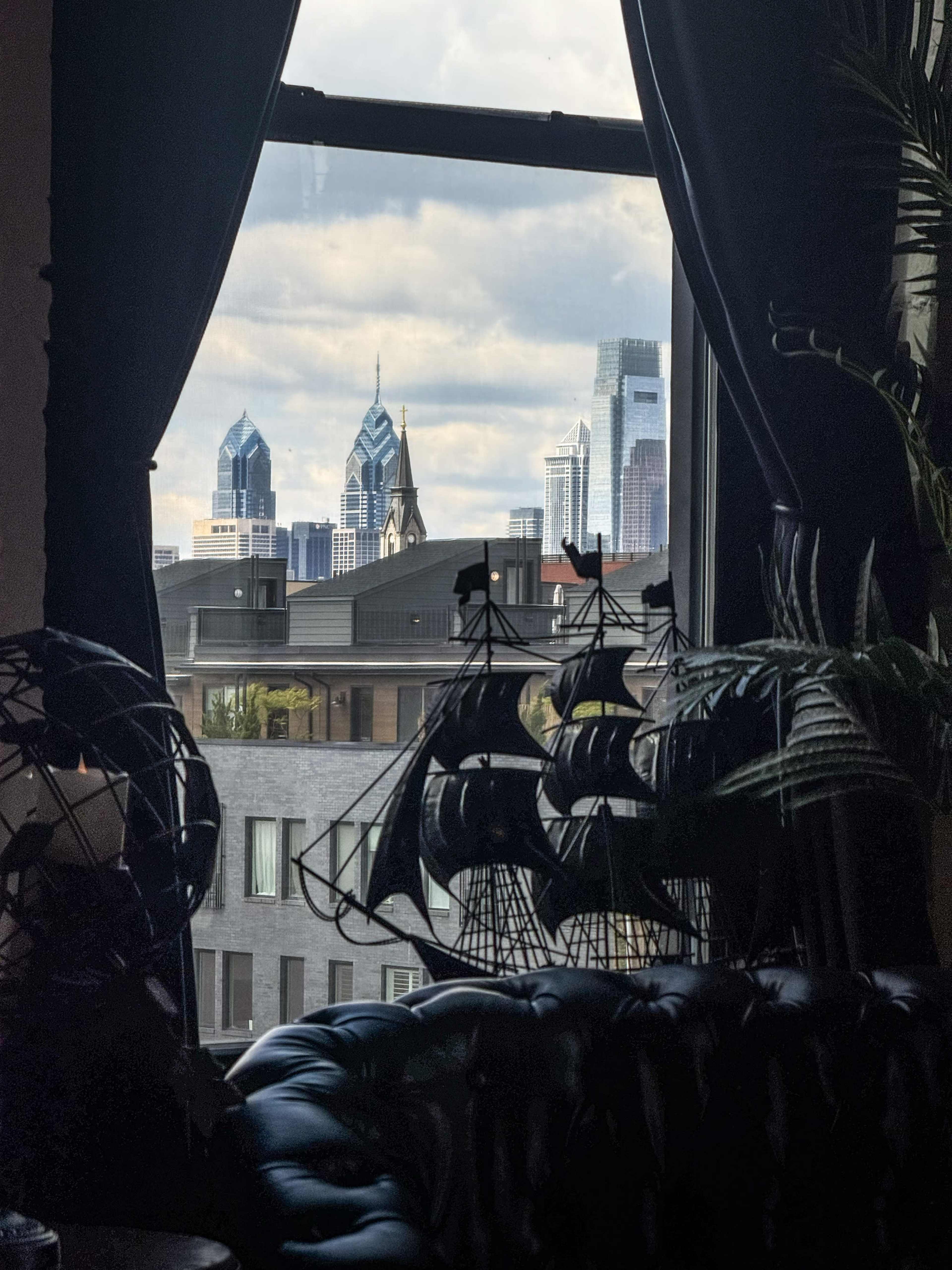 The image shows a city skyline with modern buildings visible through a window, framed by dark curtains and decorative ship models in the foreground.