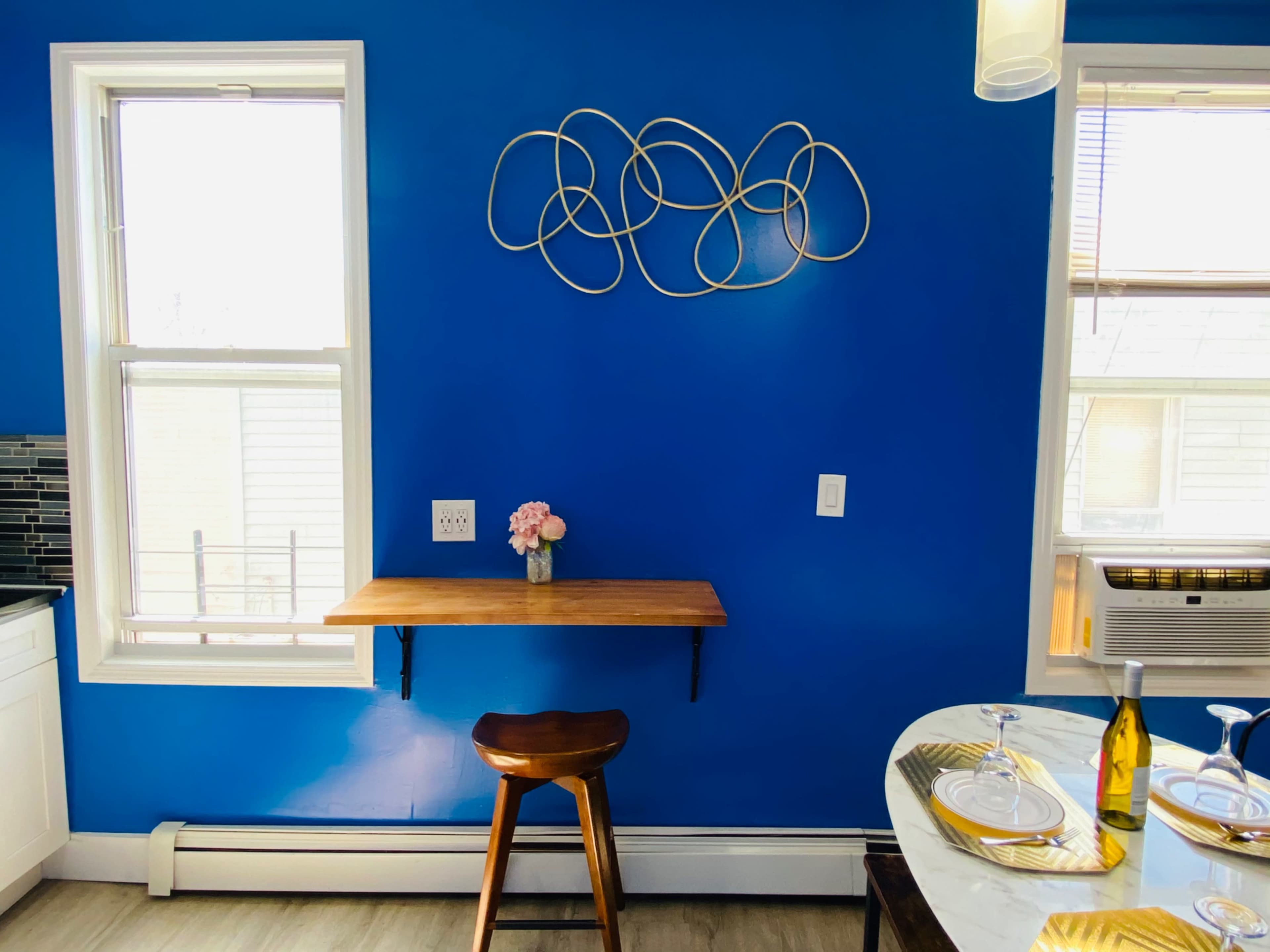 A small wooden table attached to a bright blue wall, with a vase of flowers on the table and a bar stool in front of it.
