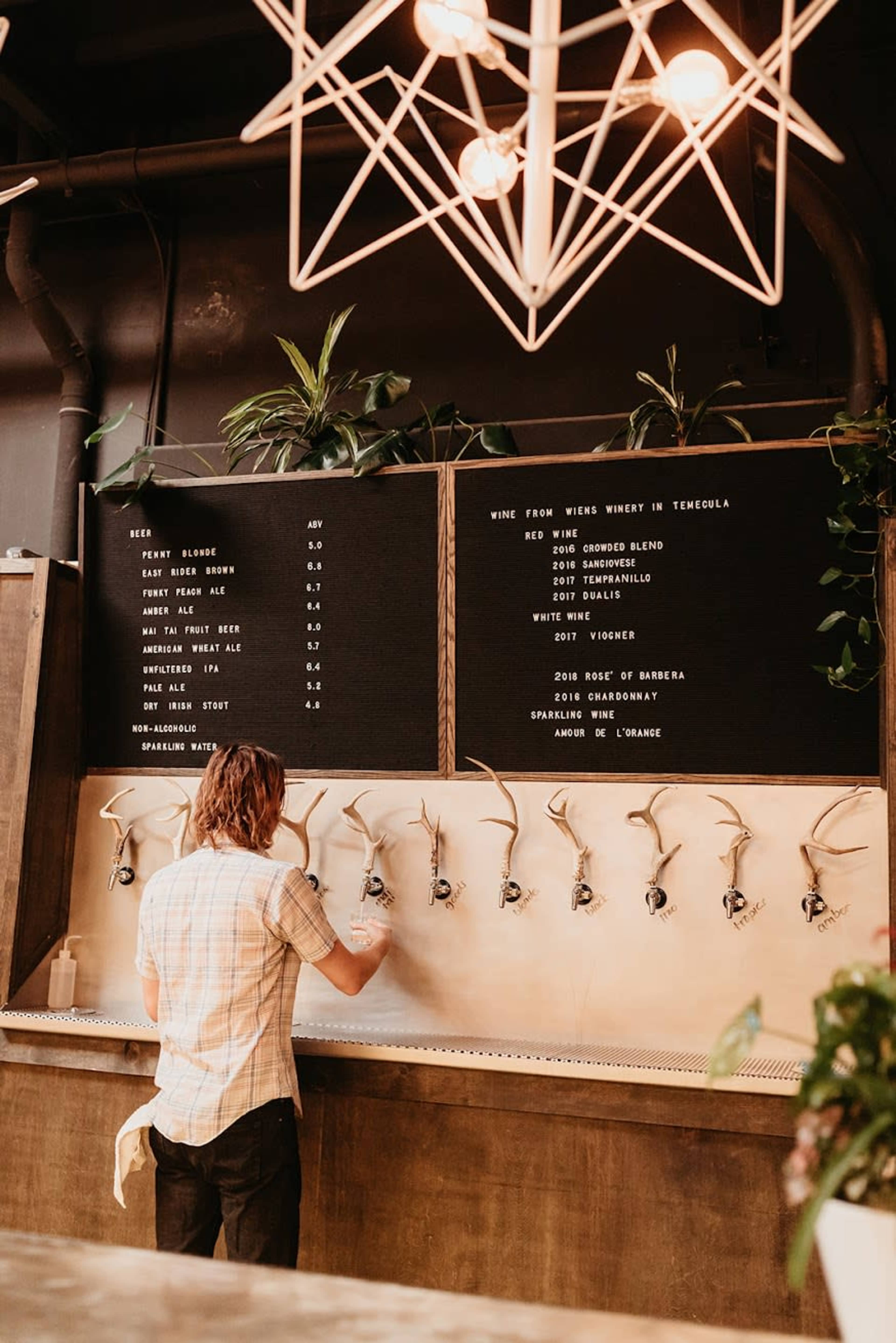A person is pouring a drink from a tap at a wine dispenser with a menu displayed above.