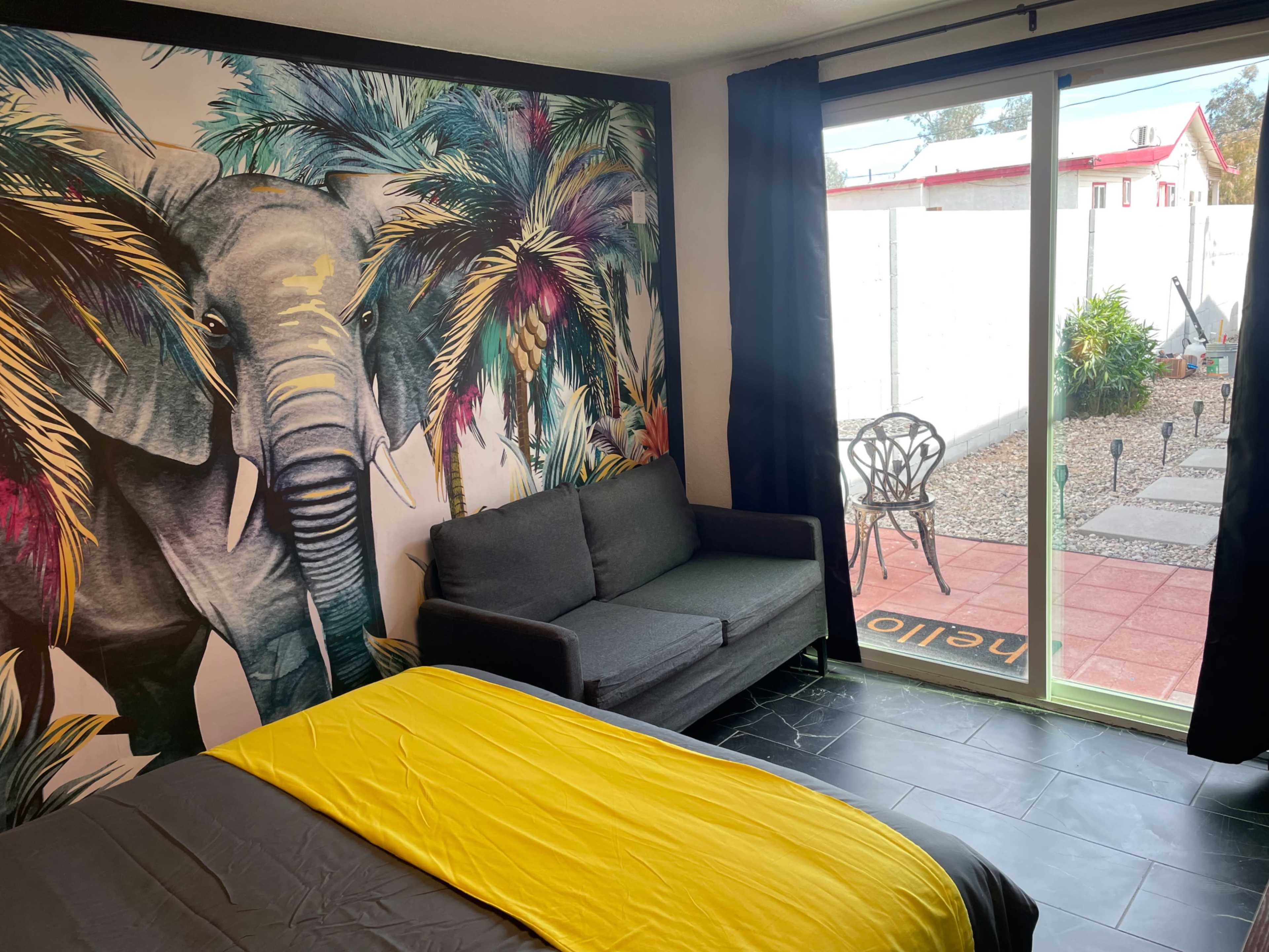 A room with a large elephant mural on one wall, a sofa, and a sliding glass door leading to a small outdoor area with a garden.