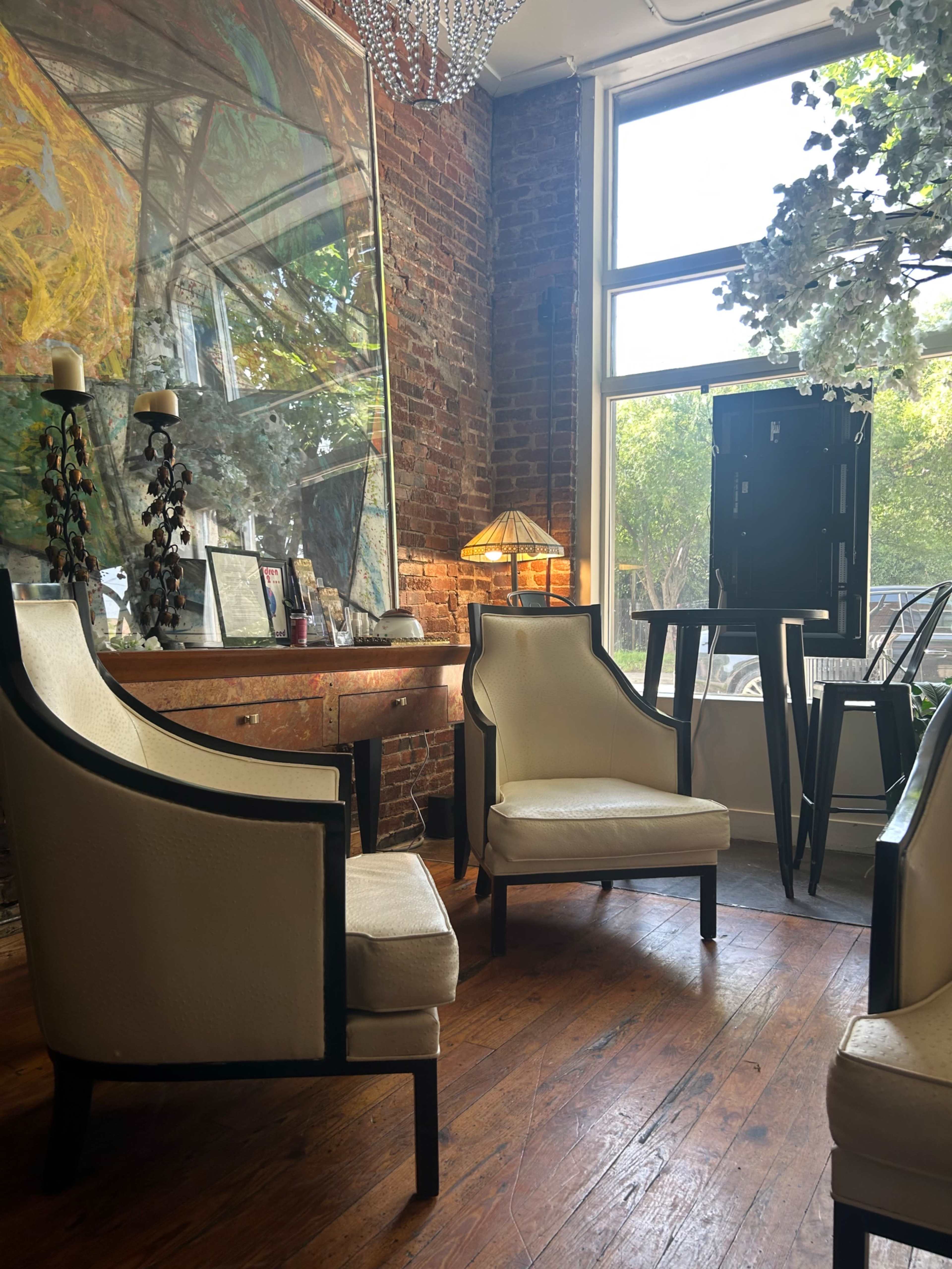 The image shows a cozy corner of a room with two white armchairs, a small table, a lamp, and a large window with sunlight streaming in.