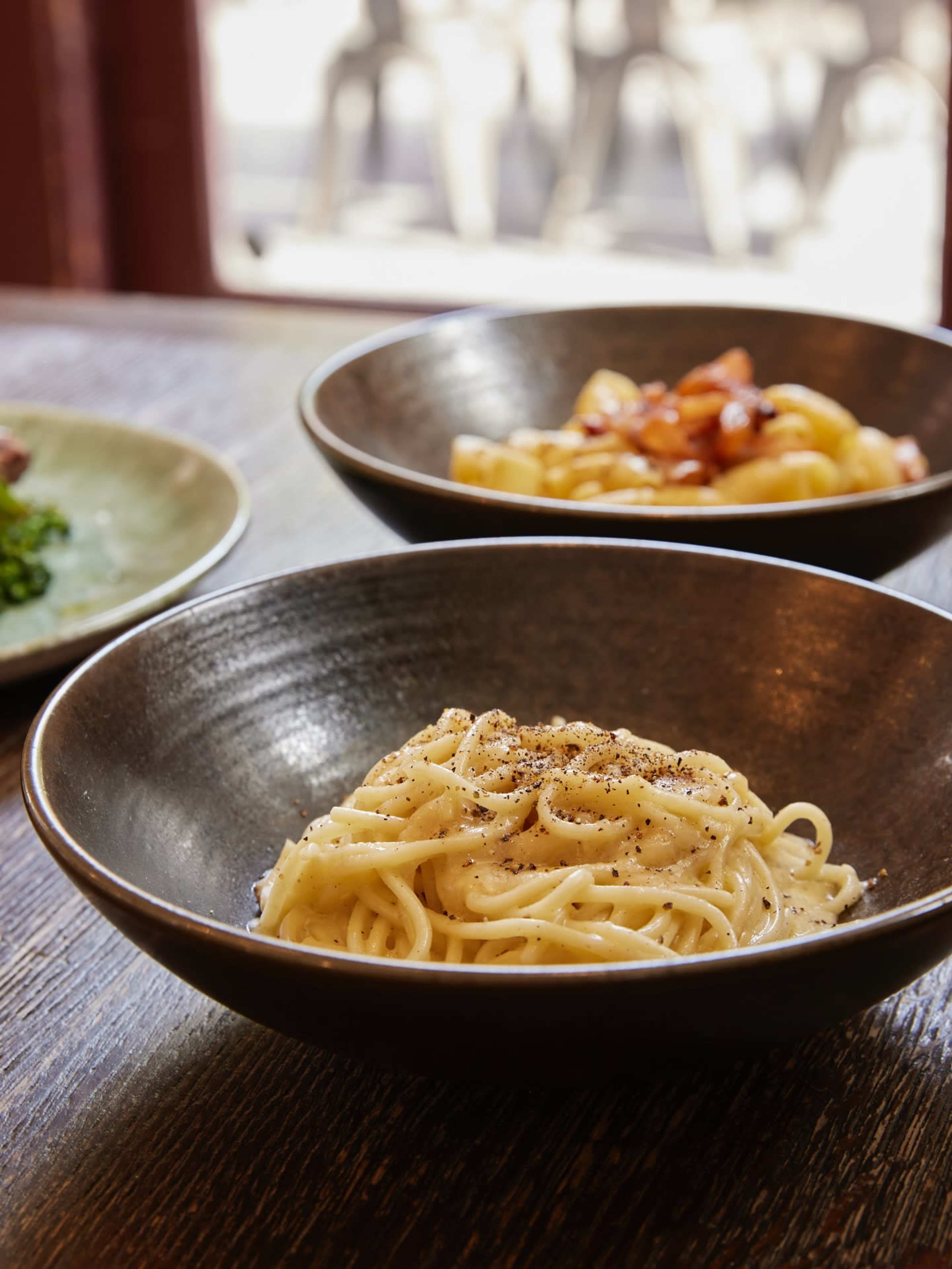 A bowl of spaghetti is topped with black pepper, seated on a wooden table with two other bowls of pasta in the background.
