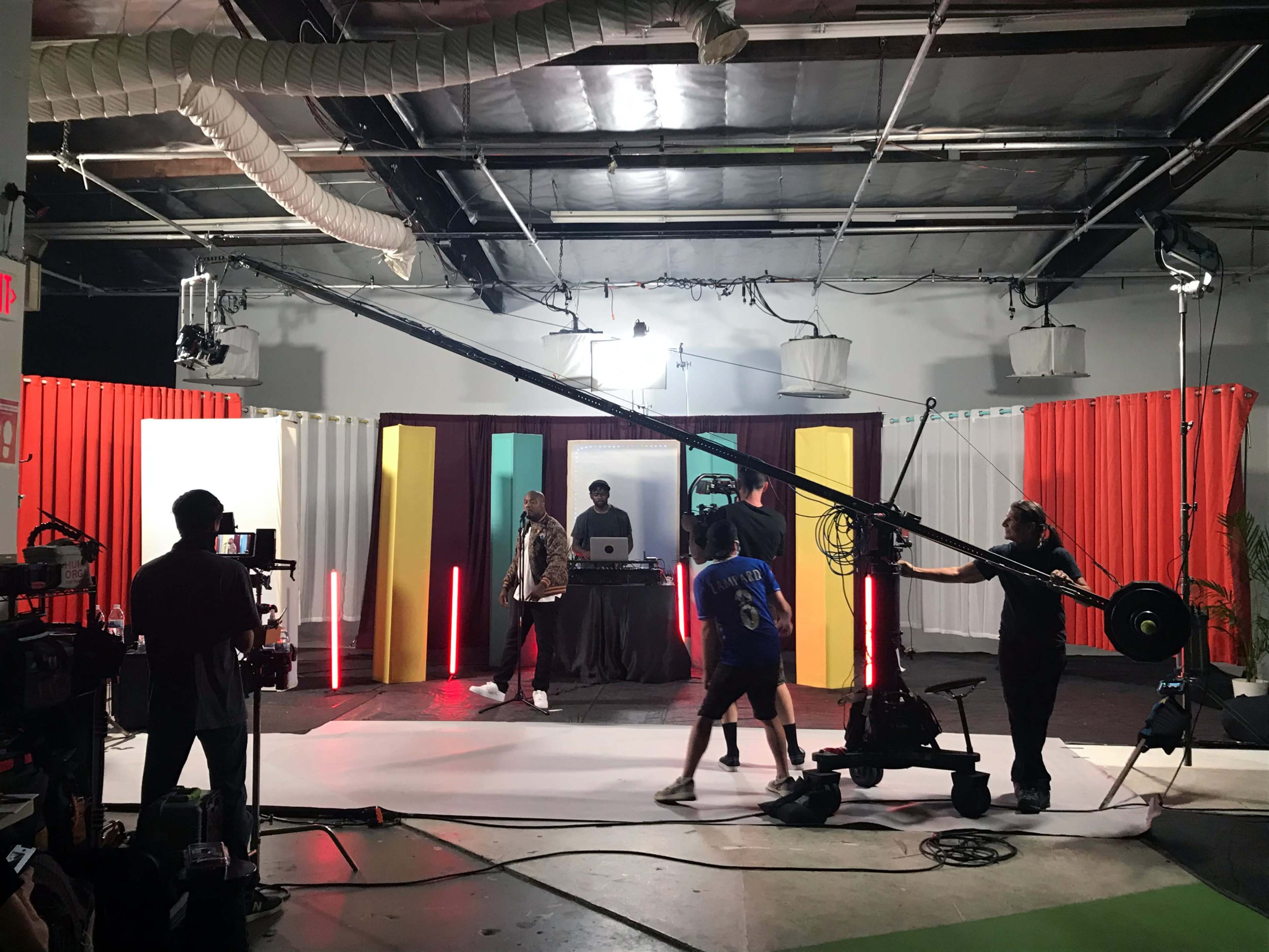 A film crew sets up equipment on a colorful studio set with a central stage for a live performance.