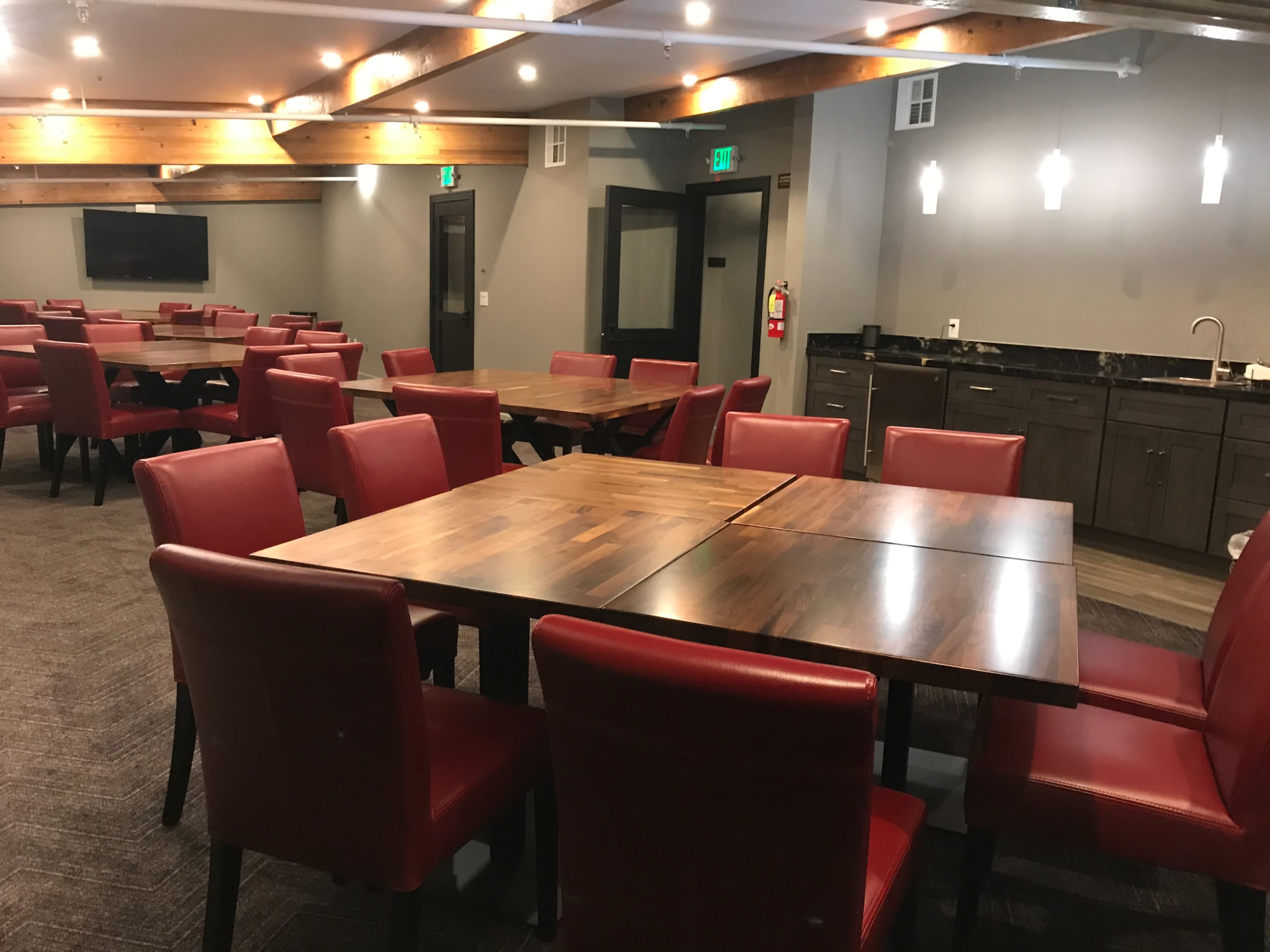 A dining area with several wooden tables surrounded by red chairs, along with a kitchenette area in the background.