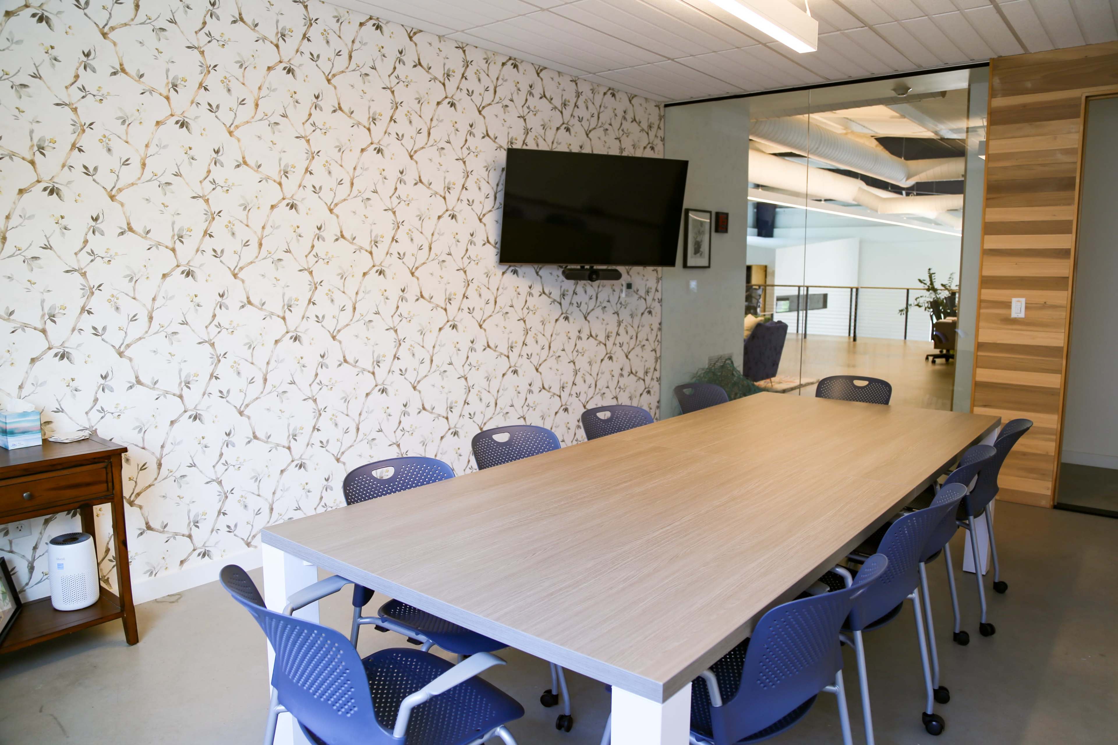 A modern conference room features a long wooden table surrounded by blue chairs, with a floral wallpaper accent and a television mounted on the wall.