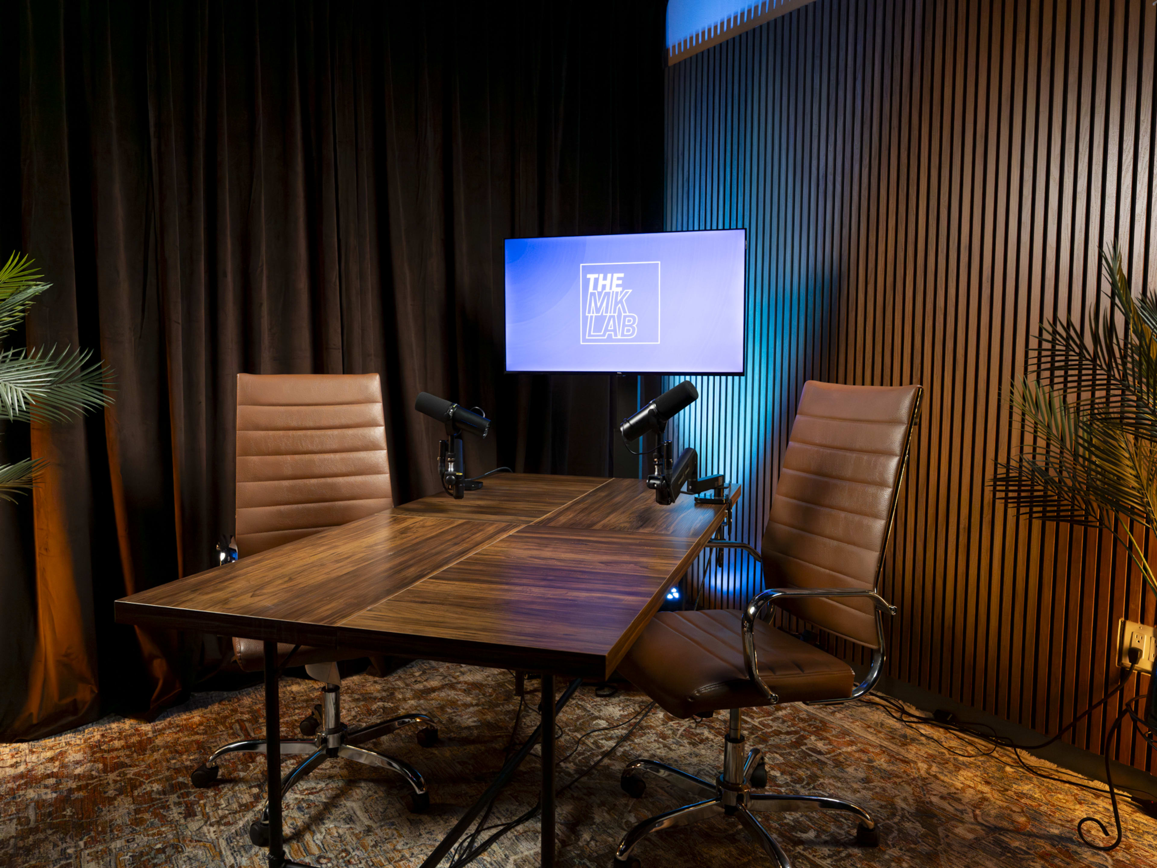 The image shows a minimalist podcast studio with two leather chairs, a wooden table, and a television screen displaying "THE WIKI LAB," set against a backdrop of dark curtains and ambient lighting.