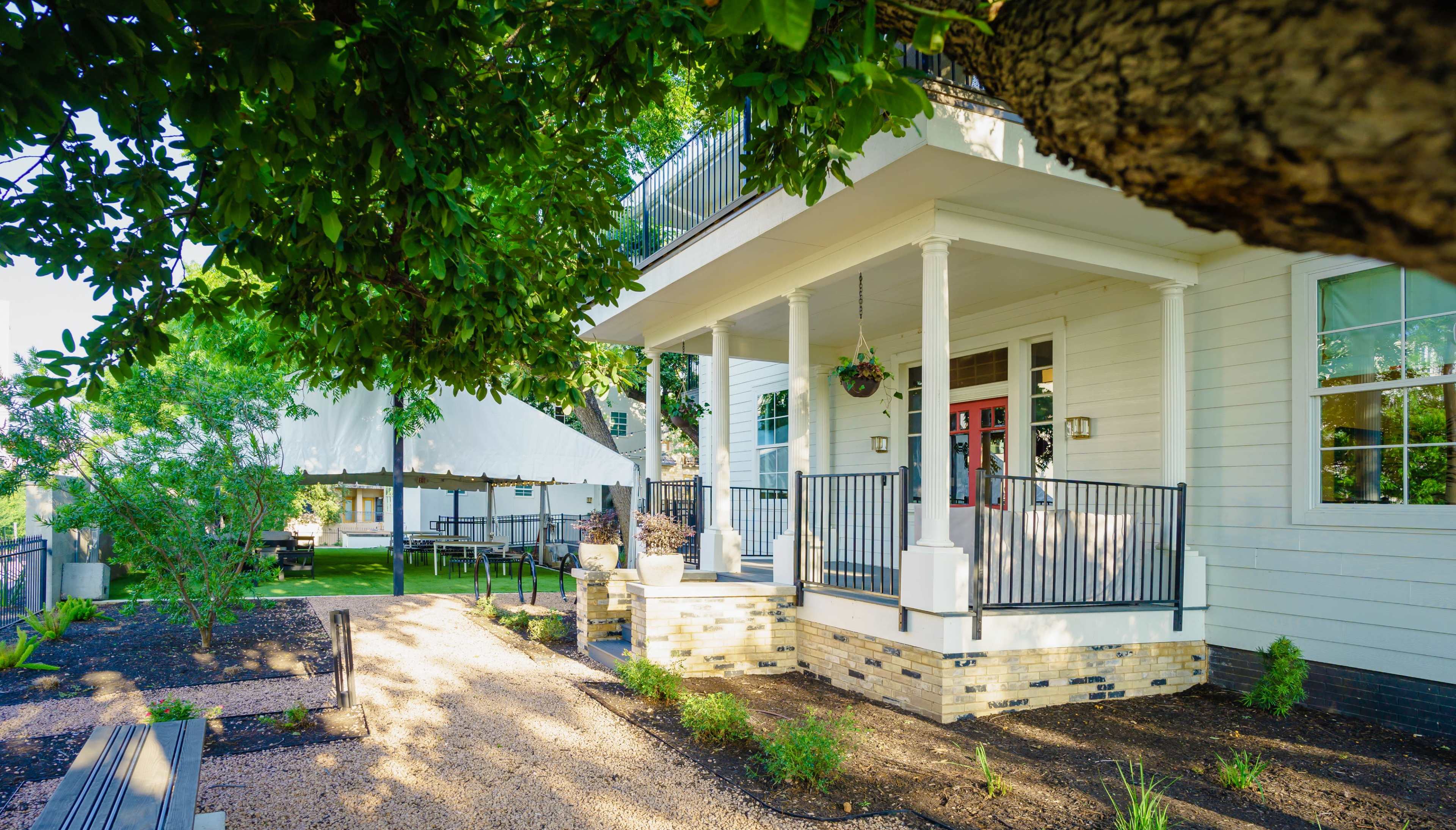 A two-story house with white siding and a red front door is situated in a landscaped garden featuring gravel pathways and a tented outdoor area.
