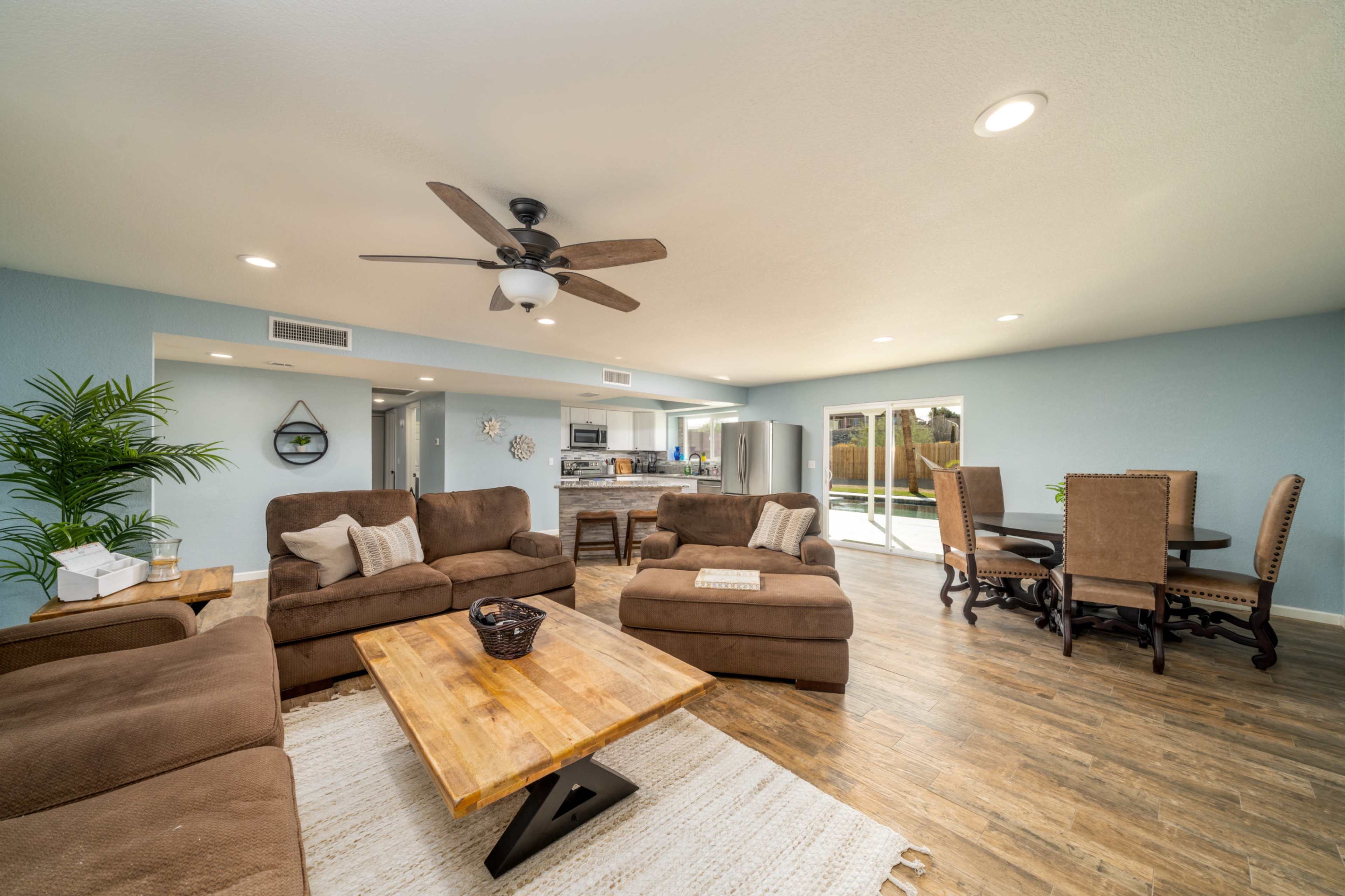 A spacious living room features brown sofas, wooden tables, a dining area, and large windows overlooking an outdoor space.