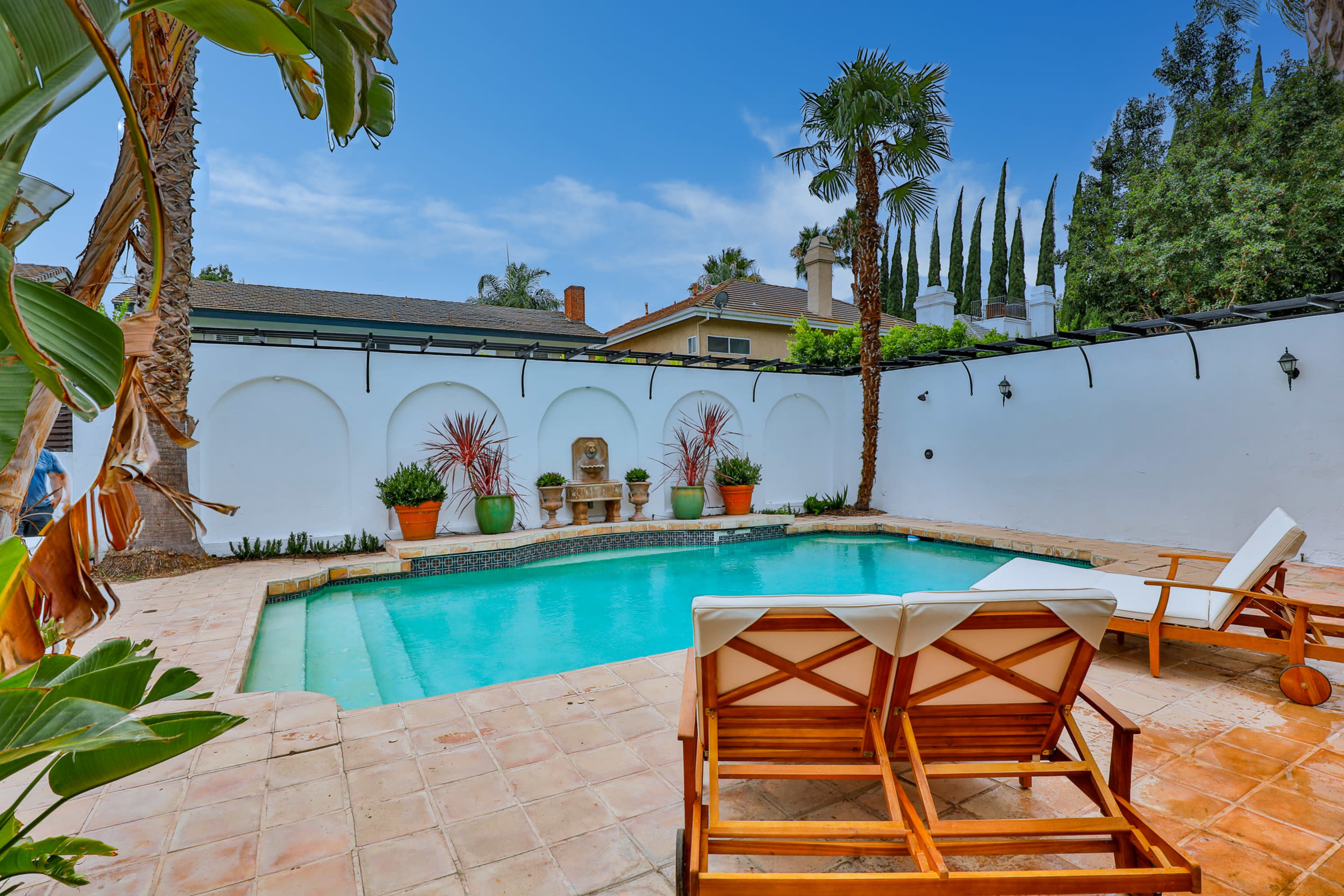 A backyard pool area featuring a turquoise swimming pool surrounded by palm trees and potted plants, with two wooden loungers positioned beside it.