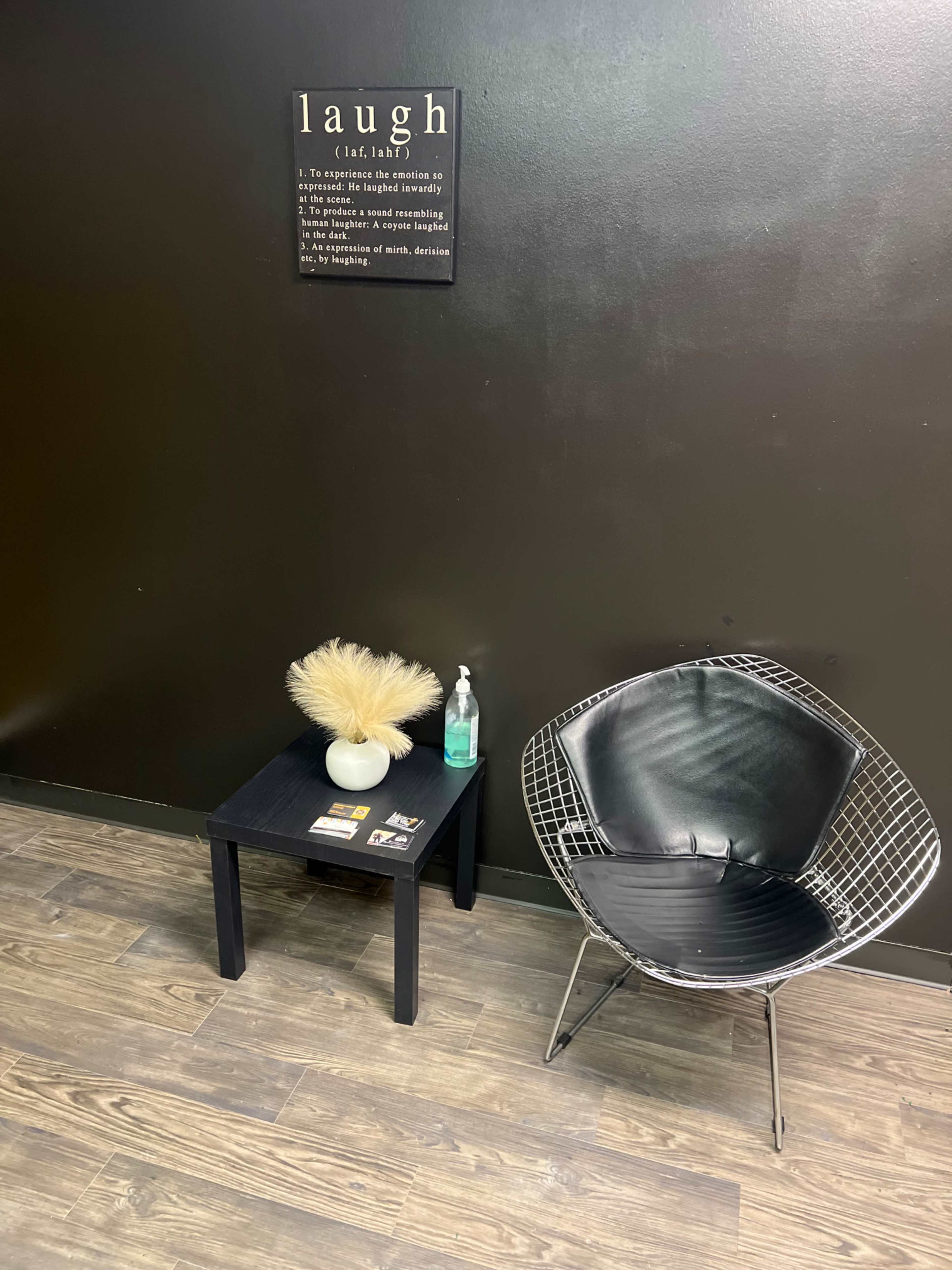 The image shows a corner of a room with a black wall, a small black table featuring a decorative plant, a hand sanitizer bottle, and a wire-framed chair.