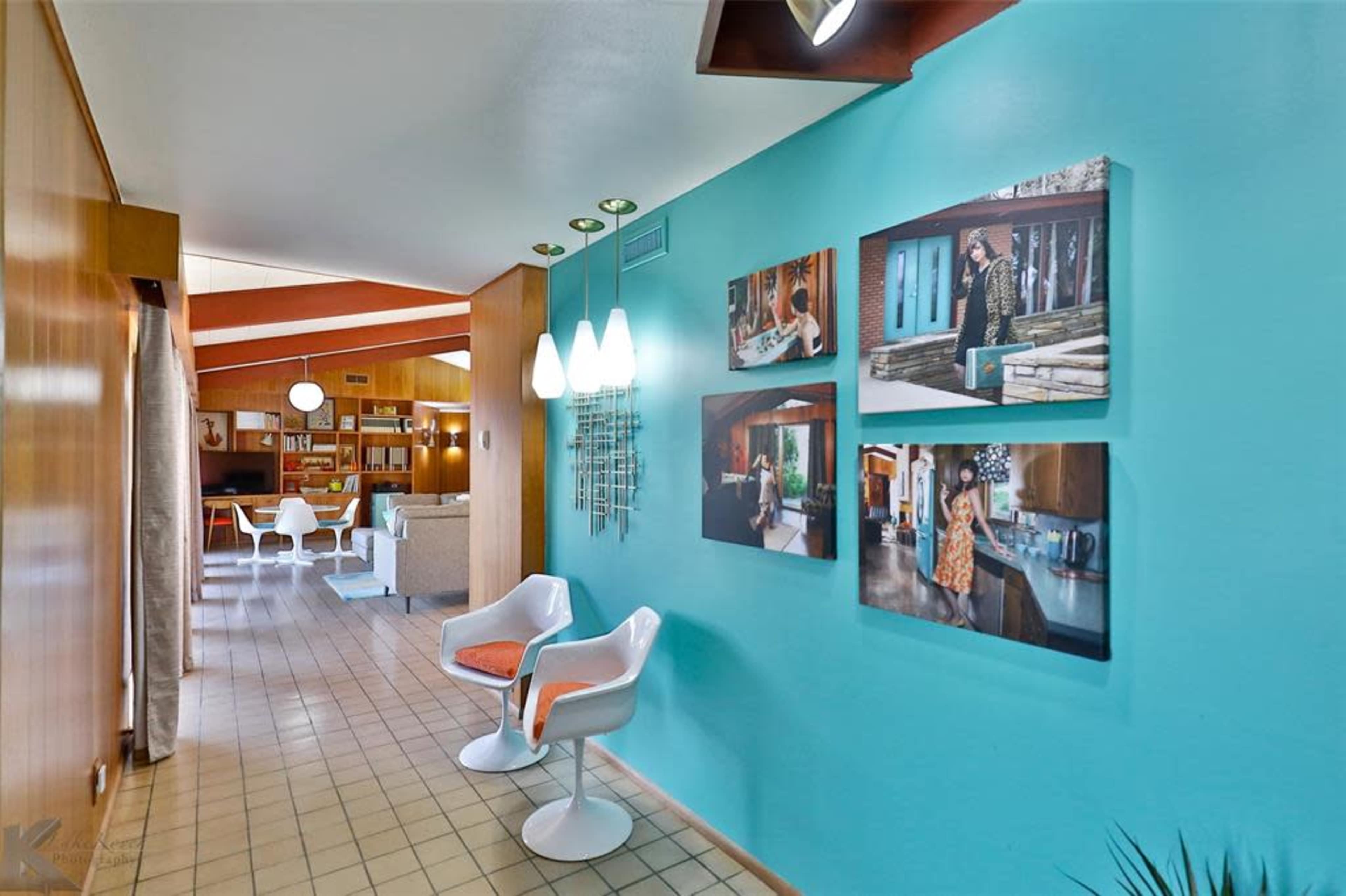 The image shows a brightly colored hallway with a blue wall featuring framed photographs, leading towards a living area with light-colored furniture.