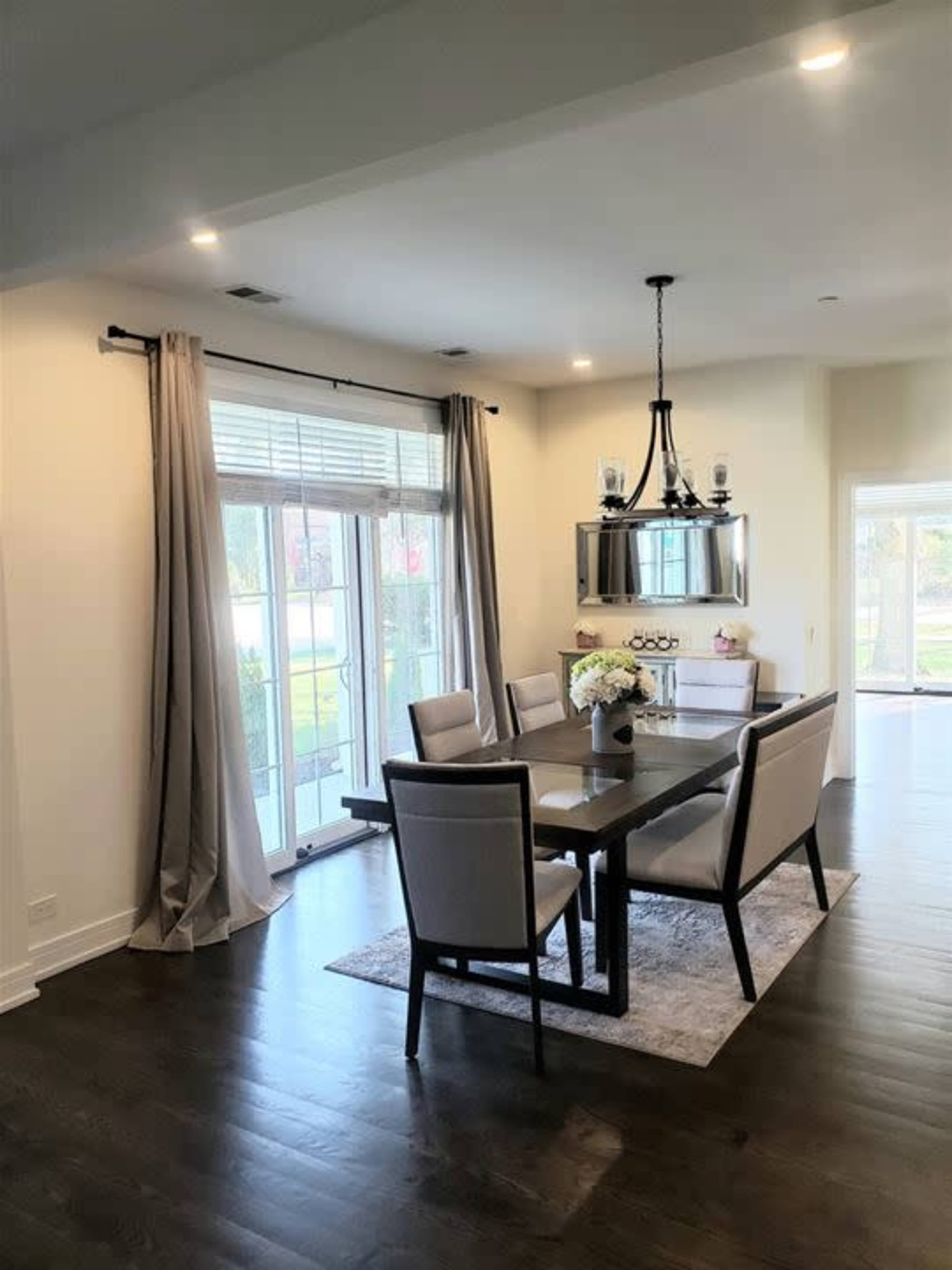 A modern dining area features a wooden table with upholstered chairs, large windows lined with curtains, and a chandelier above.