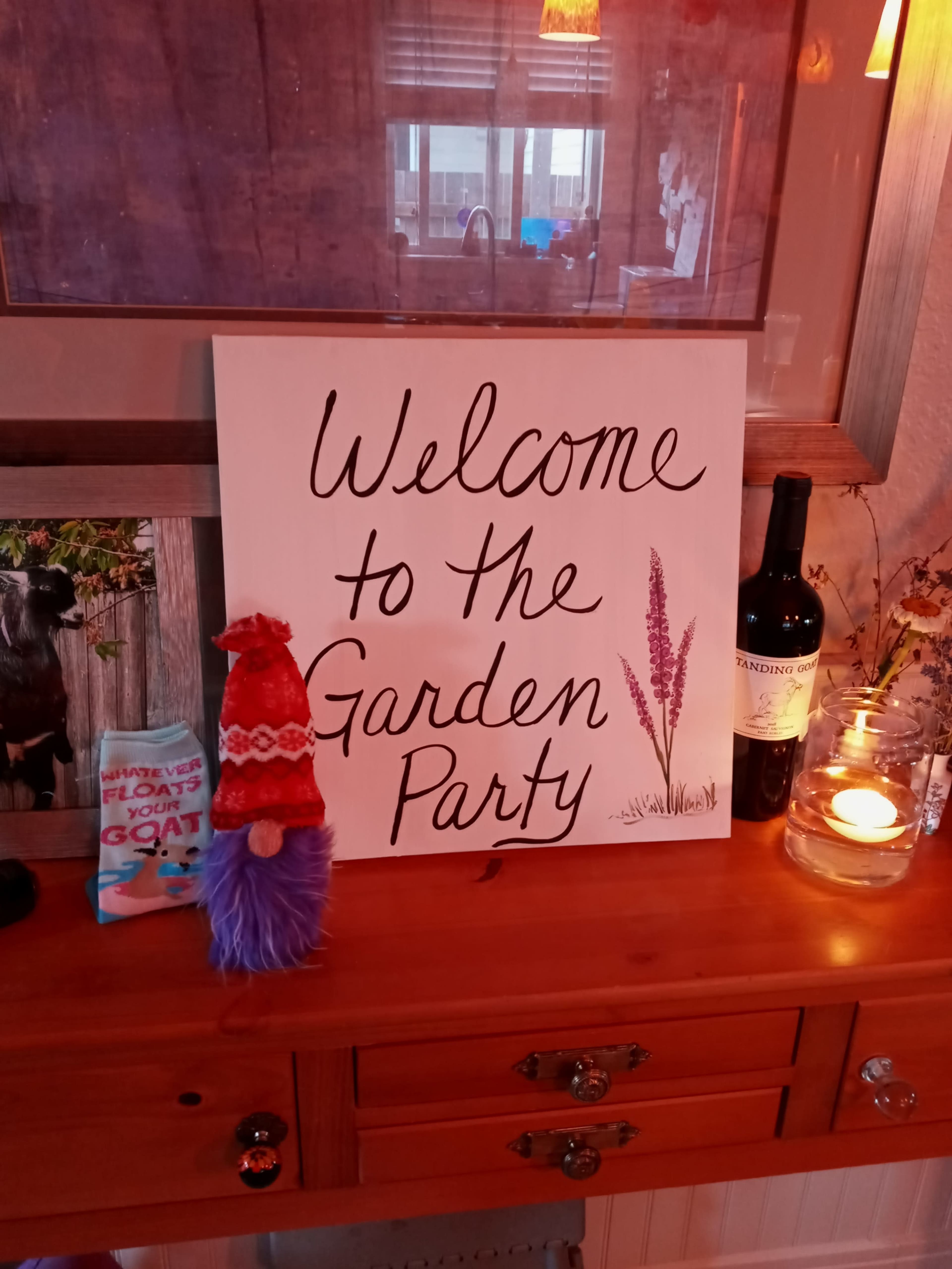 A decorated table features a welcome sign for a garden party, a bottle of wine, a candle, and a colorful gnome.