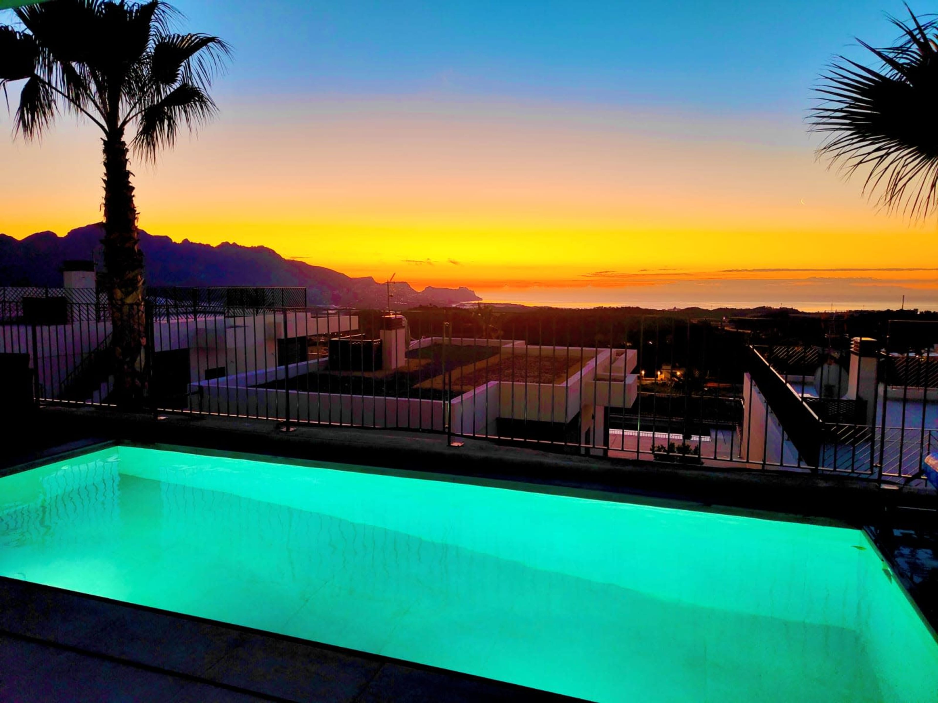 A swimming pool reflects the vibrant colors of a sunset overlooking distant mountains and the sea.