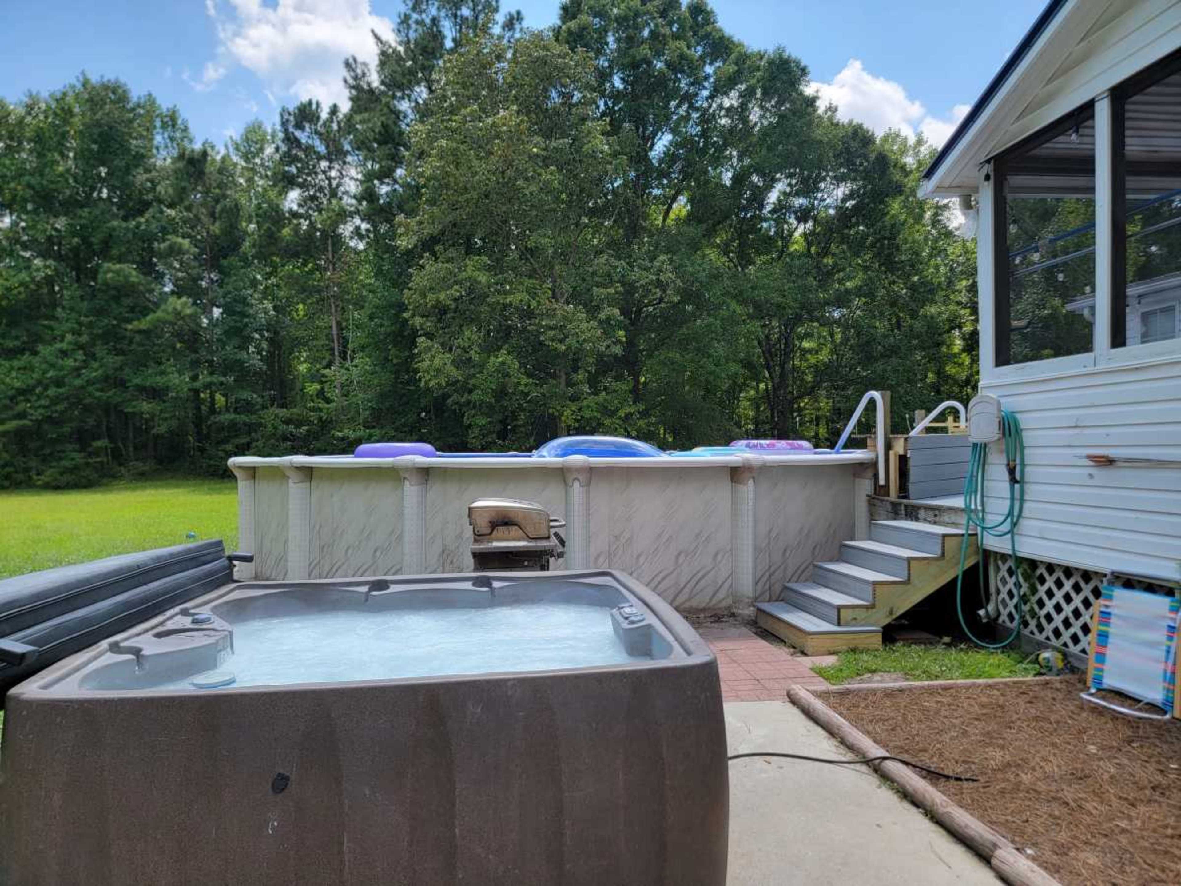 A hot tub is positioned in the foreground next to a pool surrounded by a wooden deck and stairs, with a grassy area and trees in the background.