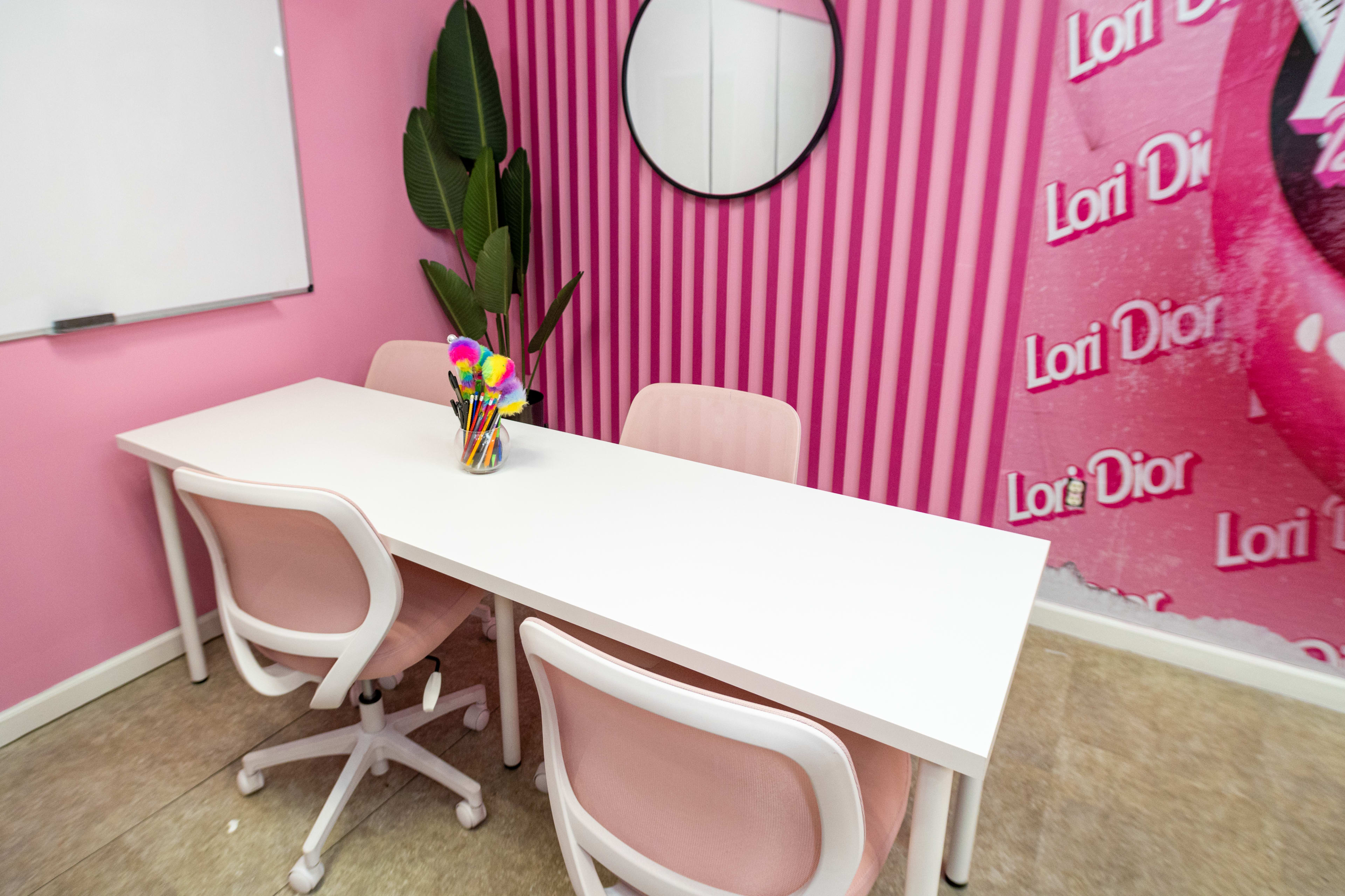 A white table with three pink chairs is positioned in a room featuring a pink striped wall and a circular mirror.