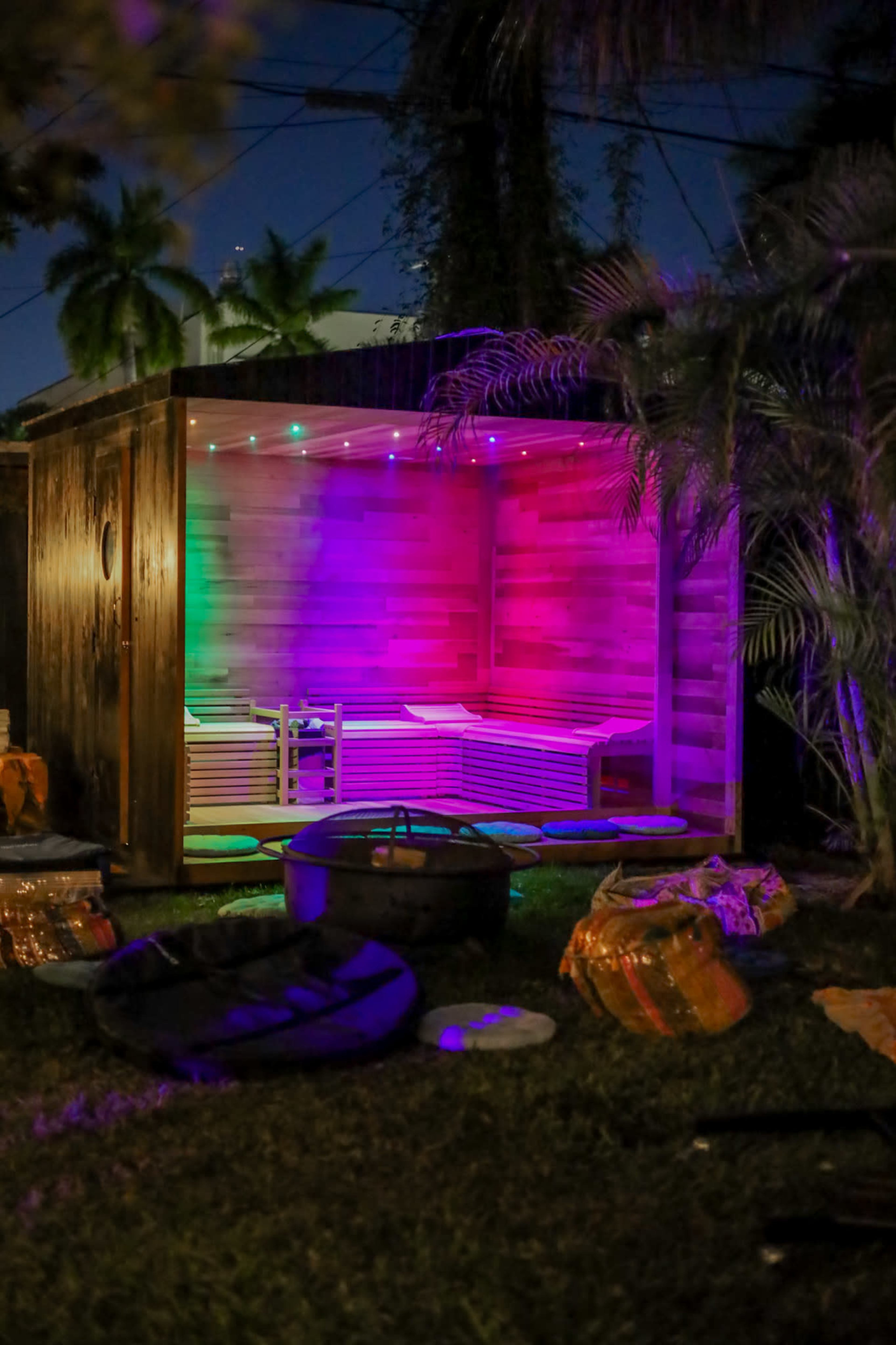 A wooden structure with colorful LED lights is situated in a yard, surrounded by lush greenery and scattered outdoor items.