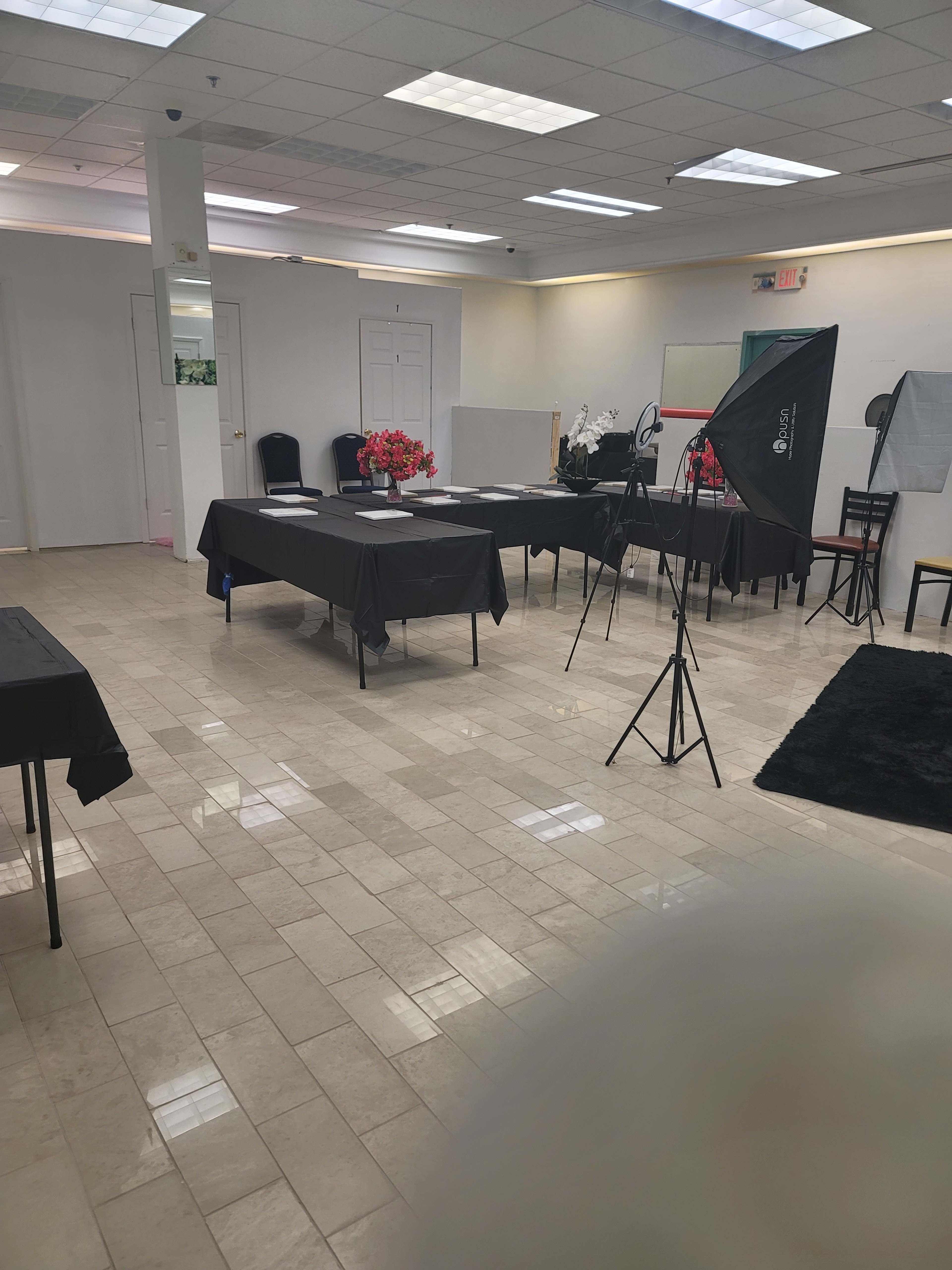 The image shows a spacious room with several tables covered in black cloth, equipped with flowers, and a lighting setup on a tripod.