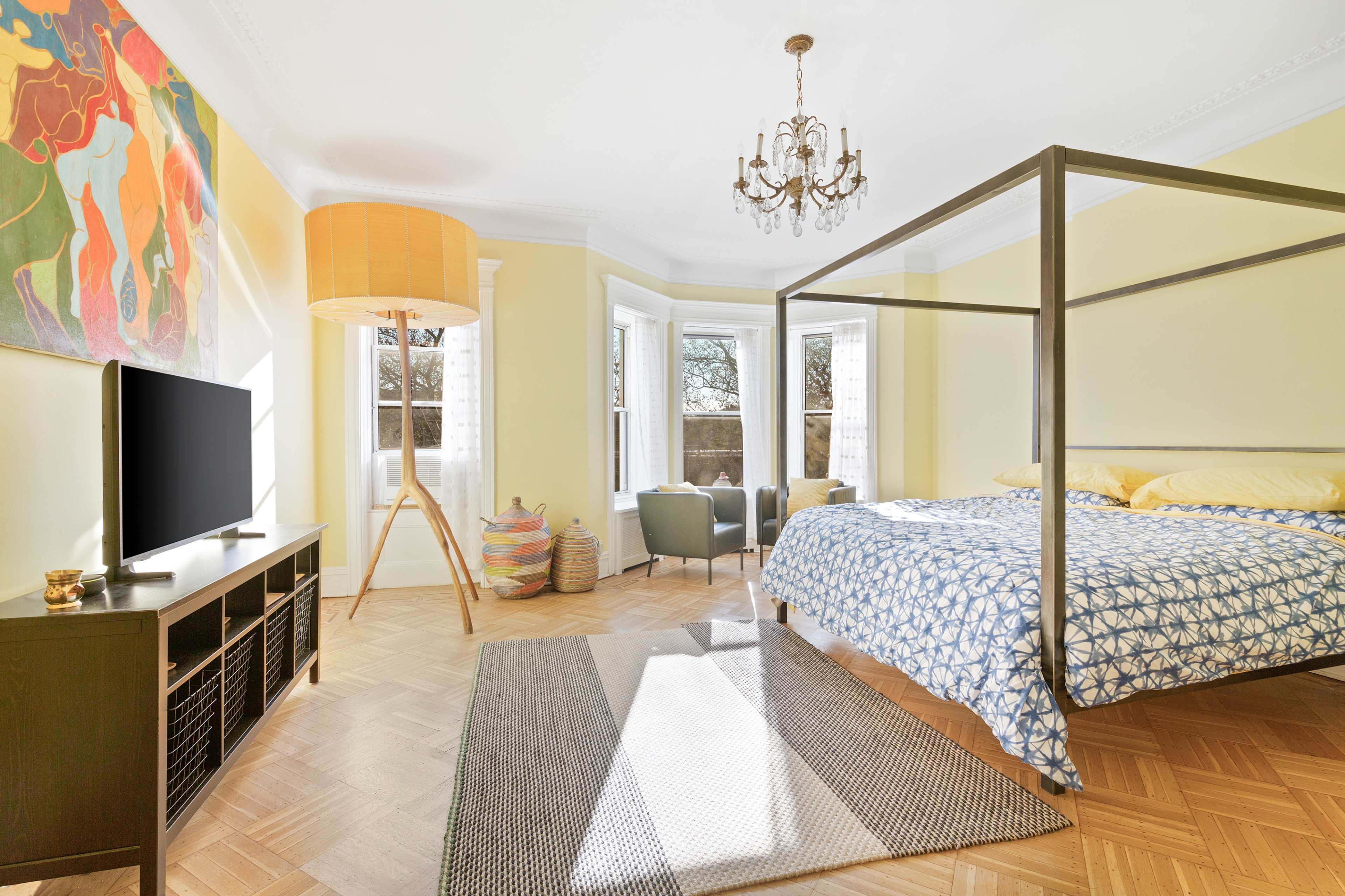 The scene depicts a bright bedroom featuring a four-poster bed with a patterned bedspread, a television on a black console, a large floor lamp, and windows that allow natural light.