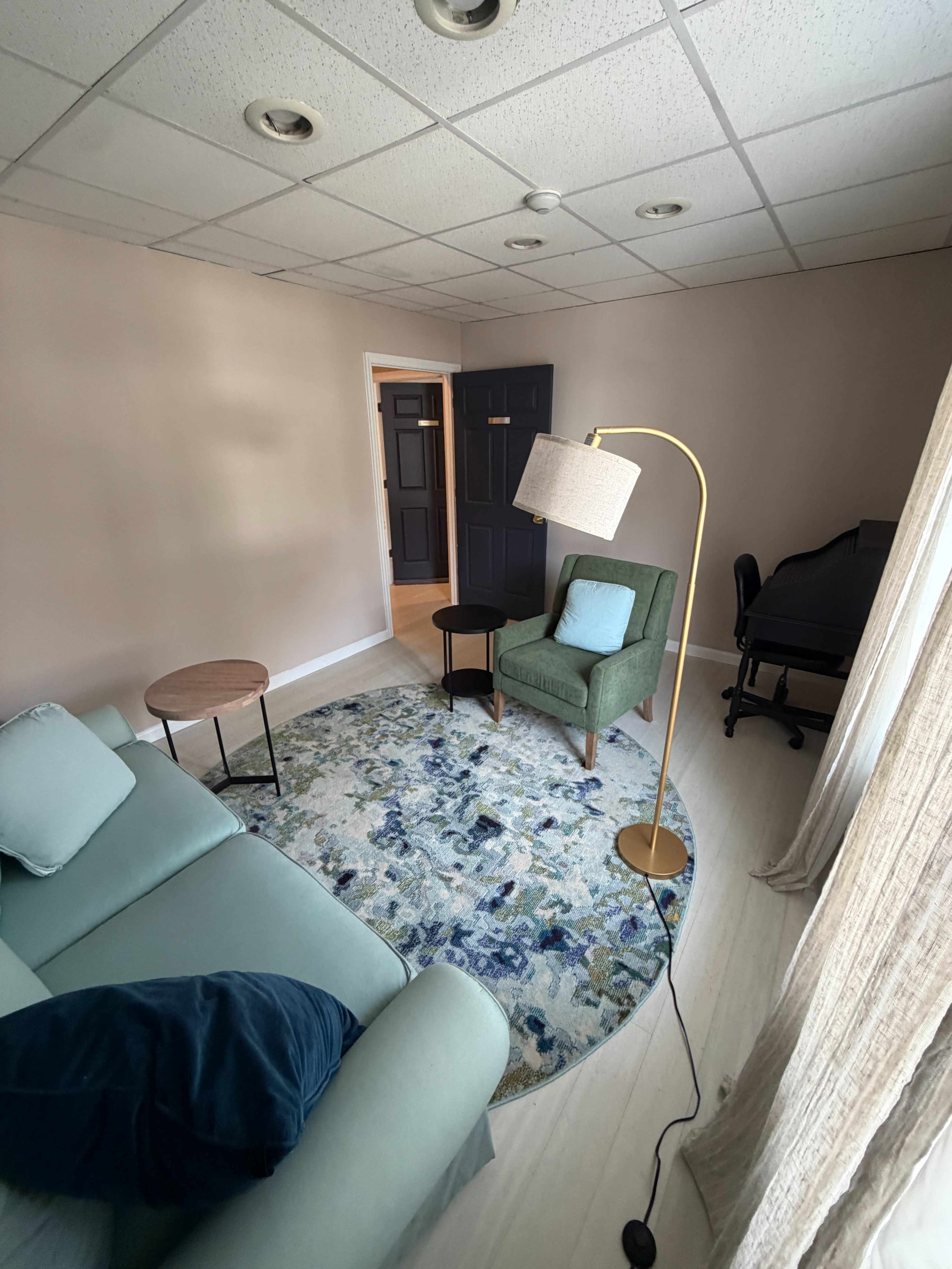 The image shows a light-colored room with a green armchair, a small round table, a lamp, and a rug featuring floral patterns, along with a doorway leading to another space.