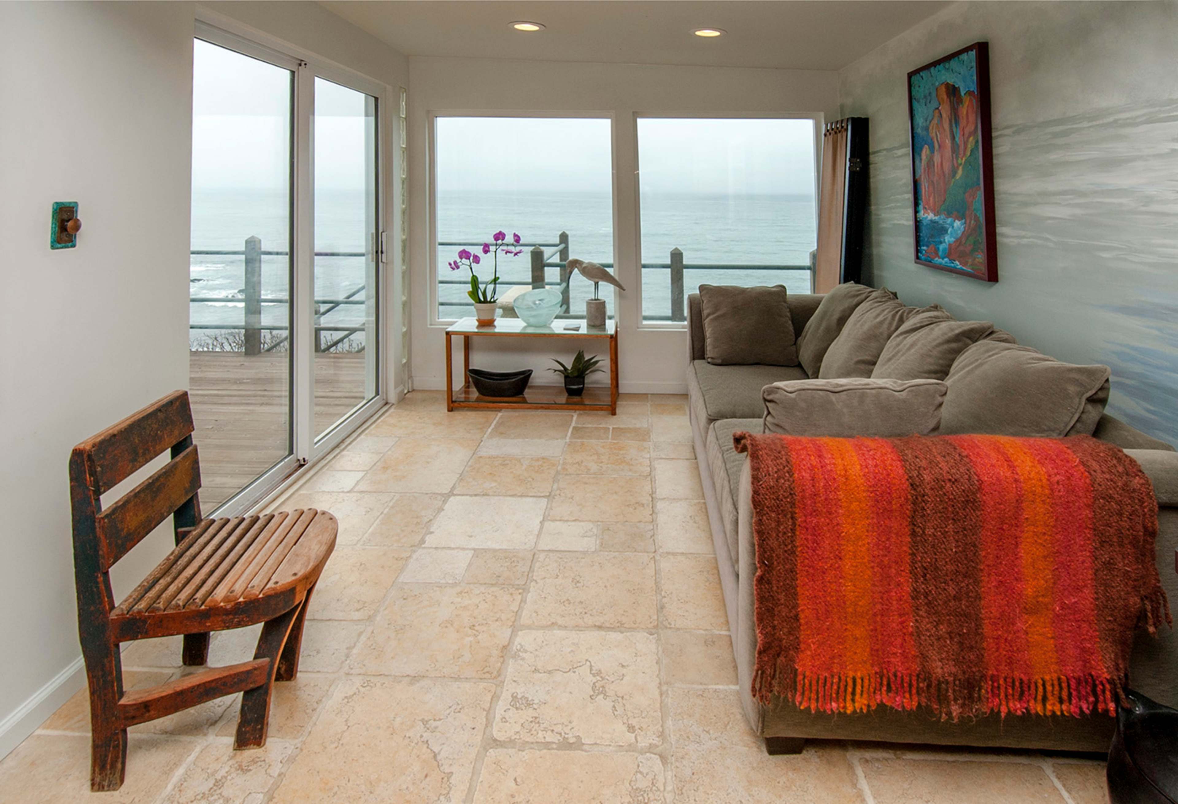 The room features a beige sofa with a red and orange throw blanket, a wooden chair, and large windows overlooking the ocean.