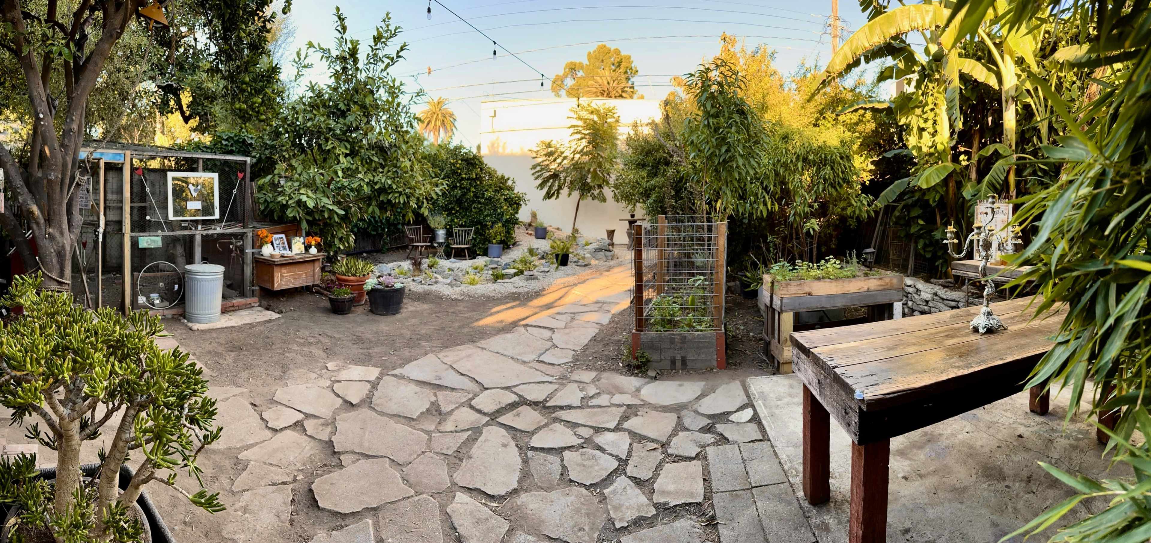 A garden with stone pathways, wooden tables, potted plants, and various planting areas surrounded by greenery.
