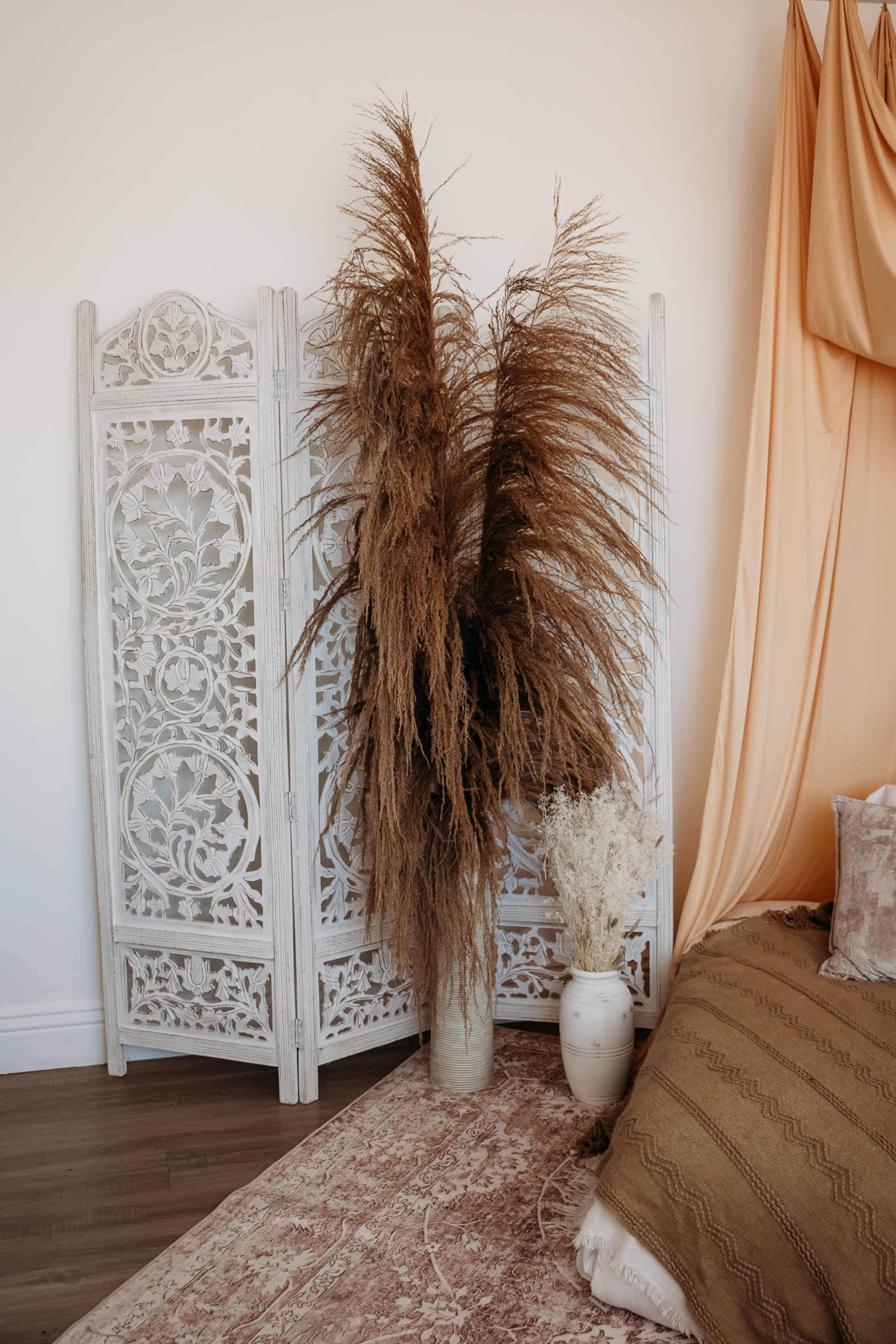 The image features a decorative room corner with a white wooden screen, tall dried plants in a vase, and a styled bed against a backdrop of draped fabric.