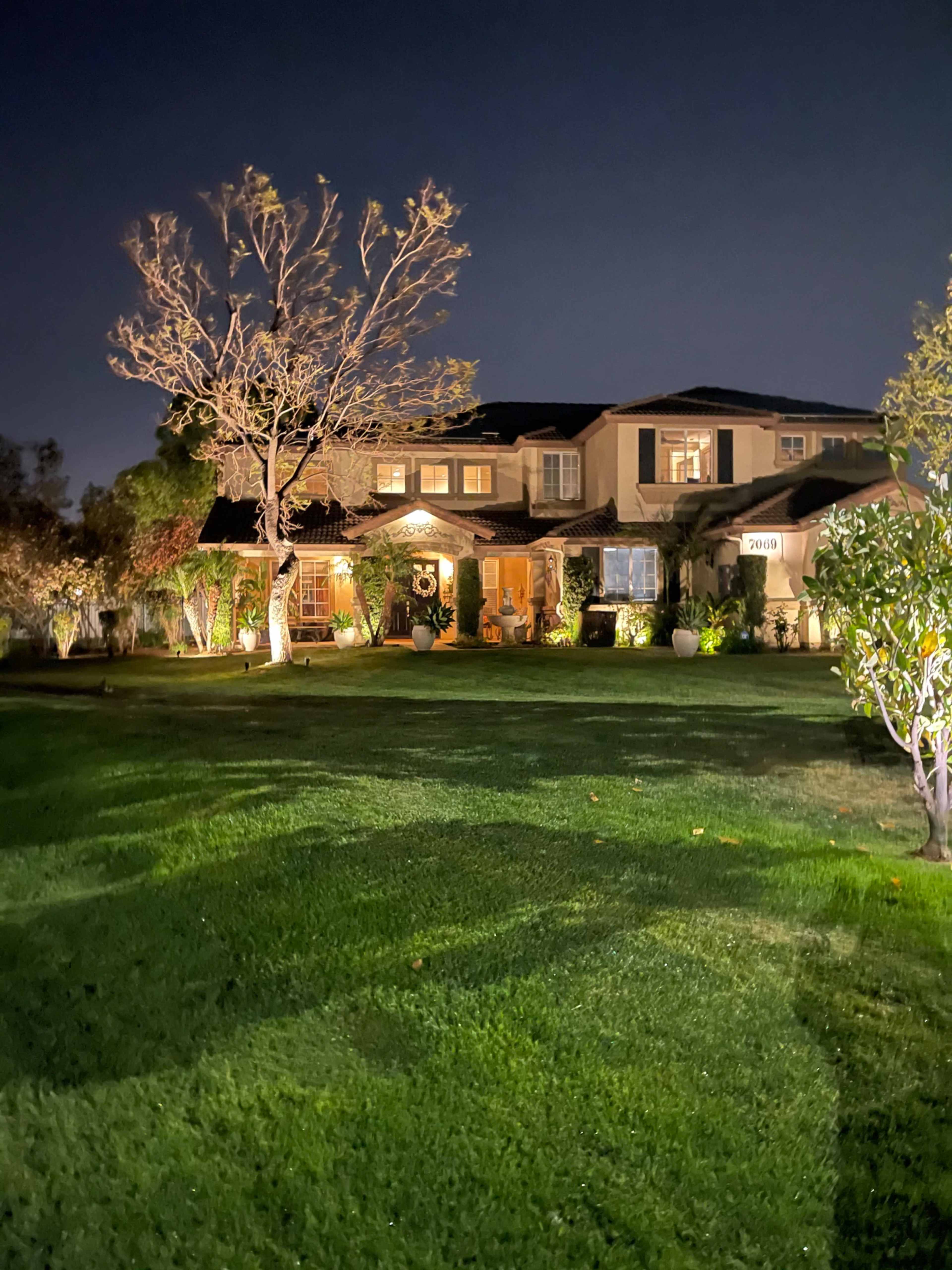 A well-lit two-story house stands in a neatly maintained lawn at night, with trees and shrubs surrounding it.