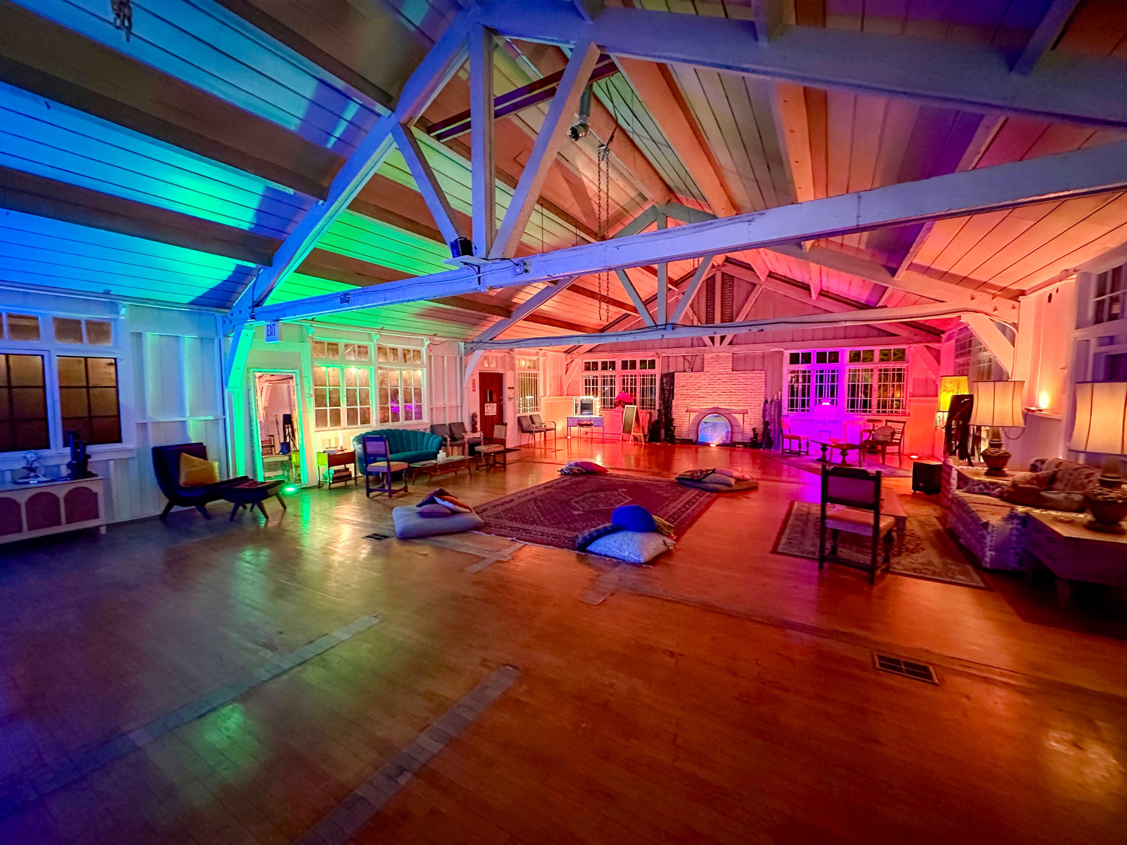 The spacious interior features high wooden beams, colorful lighting, and various seating arrangements, including cushions and chairs.