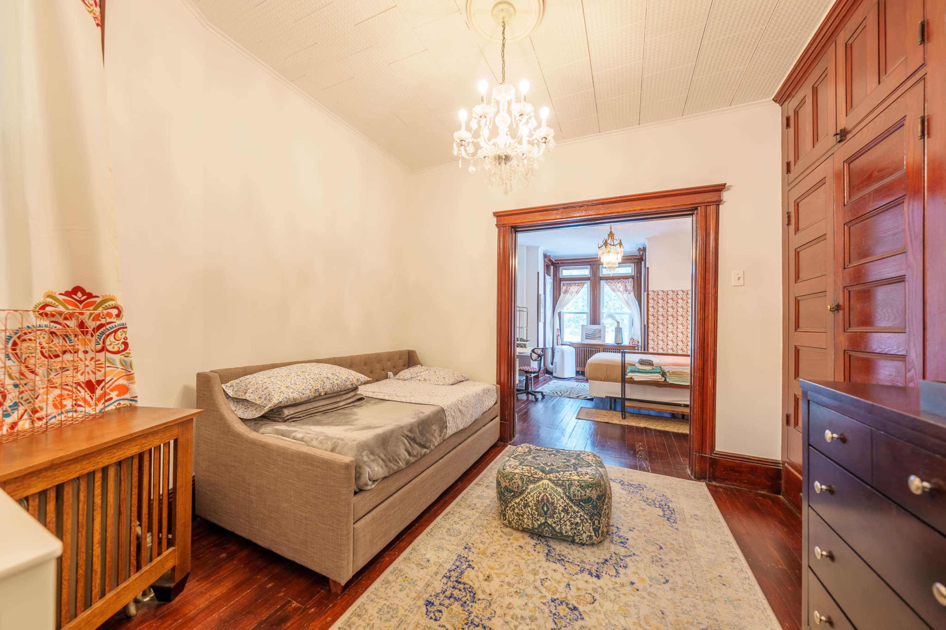 The image shows a room with a sofa bed, a patterned area rug, a decorative pouf, and a doorway leading to another room visible in the background.