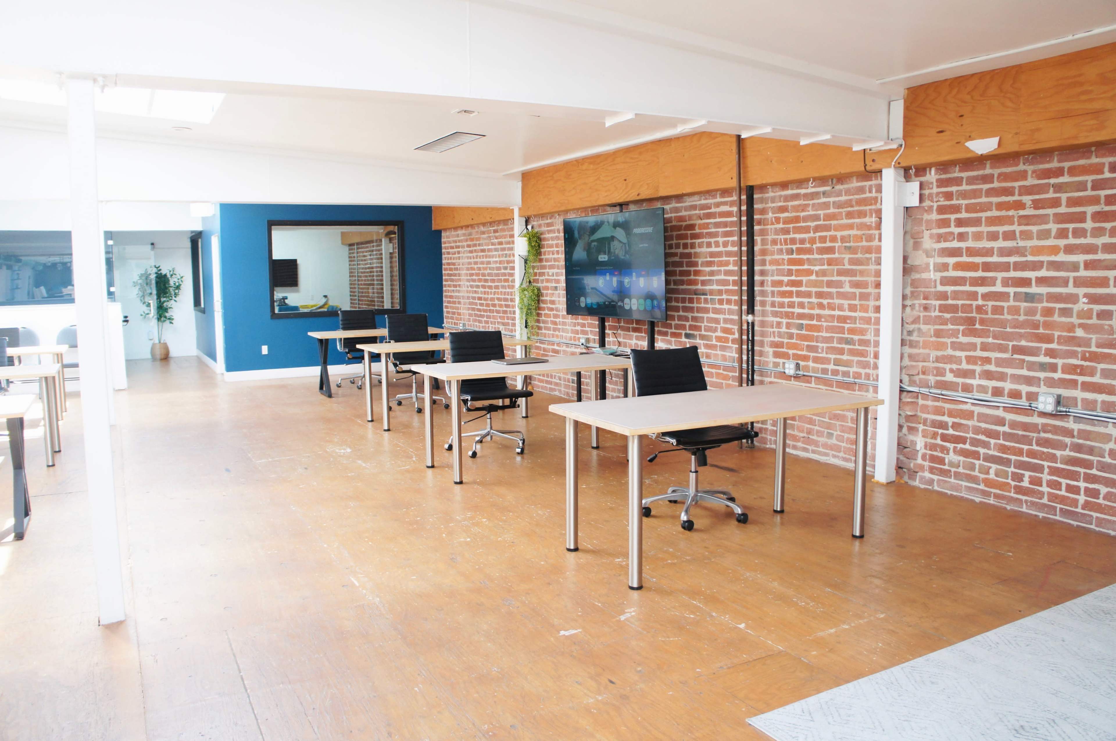 The image shows an open office space with several individual desks arranged in a row against a brick wall, featuring a large wall-mounted screen and a blue accent wall in the background.